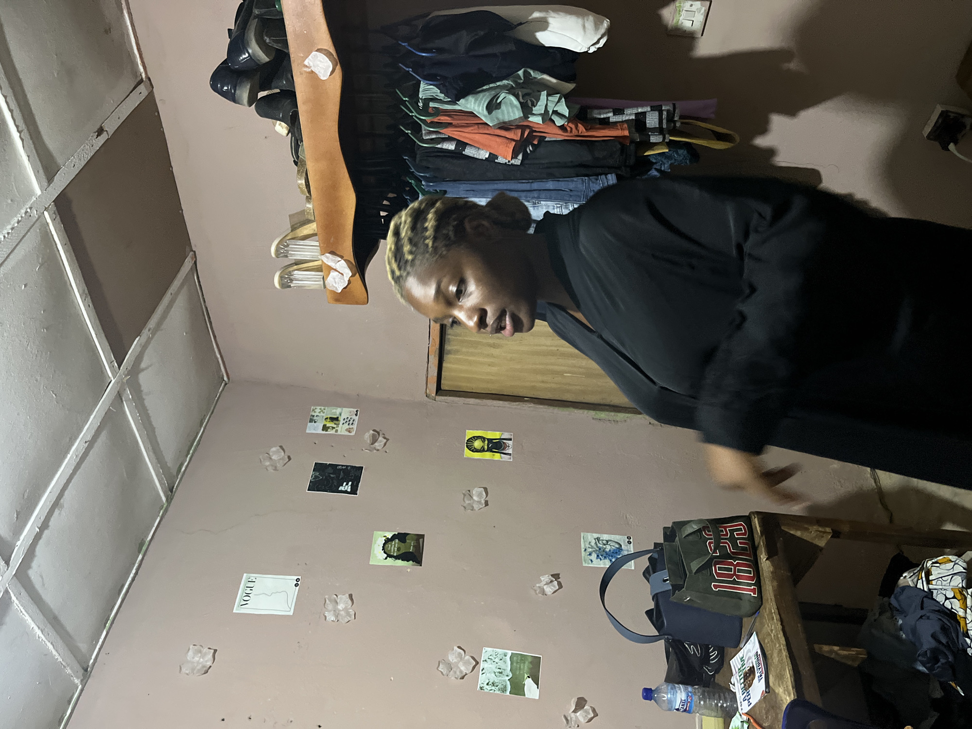 In a room that appears to be in Rafin Par, Nigeria, a young Black woman with blonde-streaked hair pulled back is standing in front of a wall adorned with several small posters and what look like decorative crystals. She is wearing a black, long-sleeved garment. To her left, a wooden table holds a blue handbag, a green tote bag with "1829" printed in red and white, and a bottle of water. Behind her, clothes hang from a rack on a wooden shelf. The room has a ceiling with visible grid lines and a wall-mounted electrical outlet with a cord plugged in.