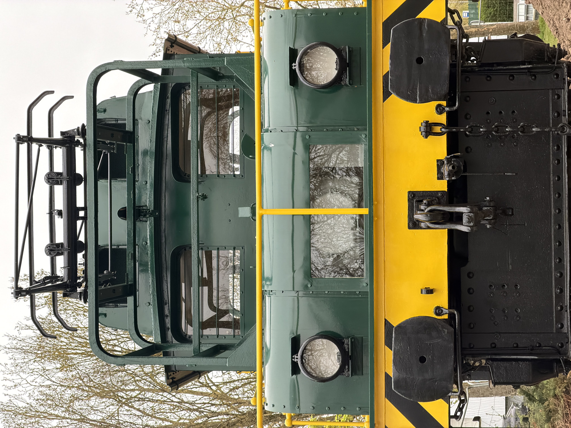 View of a green and yellow diesel locomotive on display in Esch-sur-Alzette, Luxembourg. The front of the locomotive features a prominent green cab with windows, a yellow nose with black stripes, and a black undercarriage with visible chains and couplings. Bare tree branches are visible in the background, suggesting a park or outdoor setting. The sky is overcast.