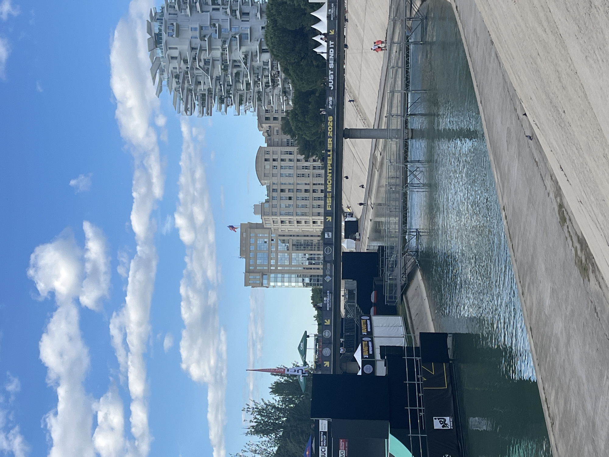 The image shows a partial view of Montpellier, France. Visible is the Le Lez River with temporary event structures along its bank, advertising "FISE Montpellier 2026," "JUST SEND IT," and "UCI World Cup." Several modern buildings are in the background, including the distinctive L'Arbre Blanc apartment building. A bridge extends over the river, with people visible on it. The sky is blue with scattered clouds. Part of a concrete wall is visible on the right side of the image.