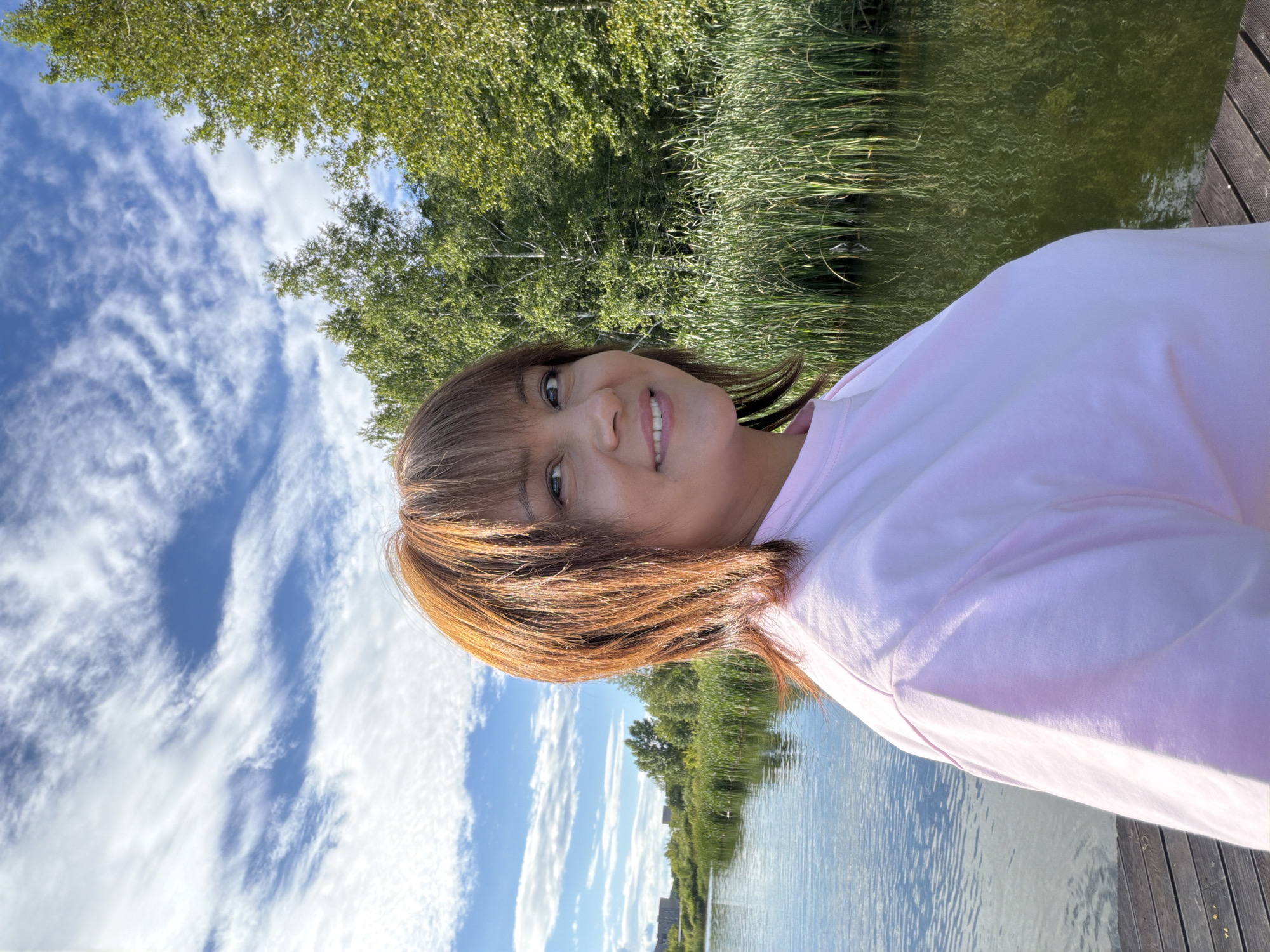 A woman with shoulder-length auburn hair takes a selfie in a natural outdoor setting. She is wearing a light pink long-sleeved shirt. The woman is smiling and looking towards the camera. Her face is partially obscured by her hair, but her eyes, nose, and mouth are visible.
The background features a body of water with tall green reeds along the edge. Beyond the reeds, trees with lush green foliage are visible. The sky above is a vibrant blue with scattered white, wispy clouds. The scene suggests it is a sunny day. Part of a wooden deck is visible in the foreground, indicating the woman might be on a pier or boardwalk. The overall mood appears cheerful and relaxed. The image is oriented vertically, with the top of the frame showing the sky and trees.