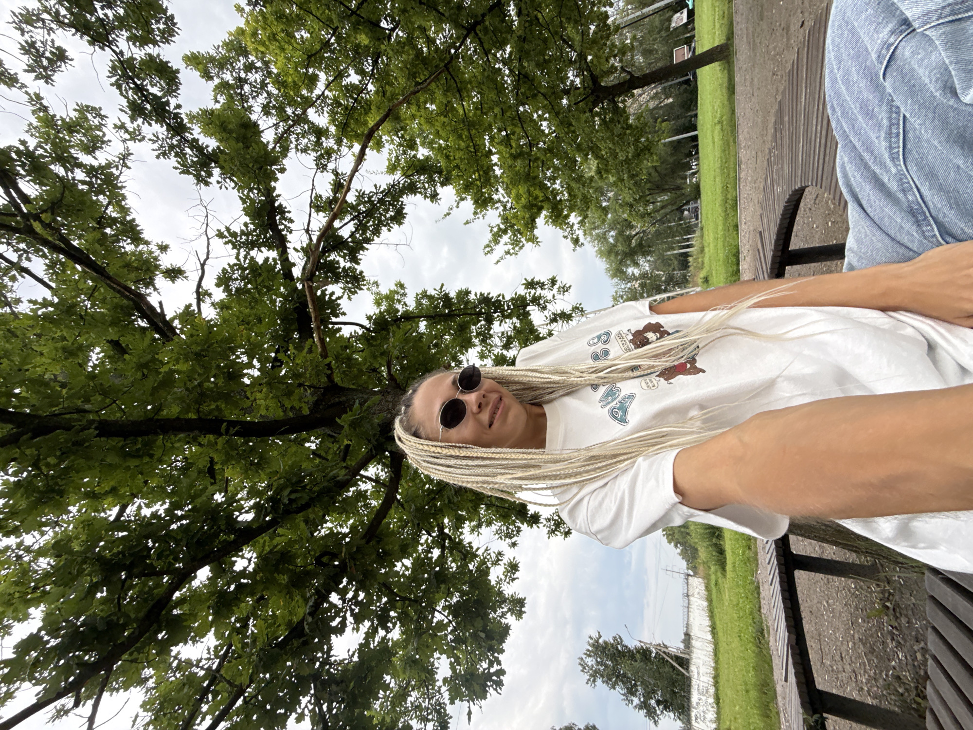 In Rusanovo, Russia, a woman with blonde braided hair and sunglasses is sitting on a wooden bench in an outdoor park. She is wearing a white t-shirt with graphics and blue jeans. The woman is holding up her phone, taking a selfie. In the background, a large tree with green leaves provides shade, and beyond that, there is a grassy field, other trees, a fence, and a cloudy sky. The overall scene suggests a casual outdoor setting.