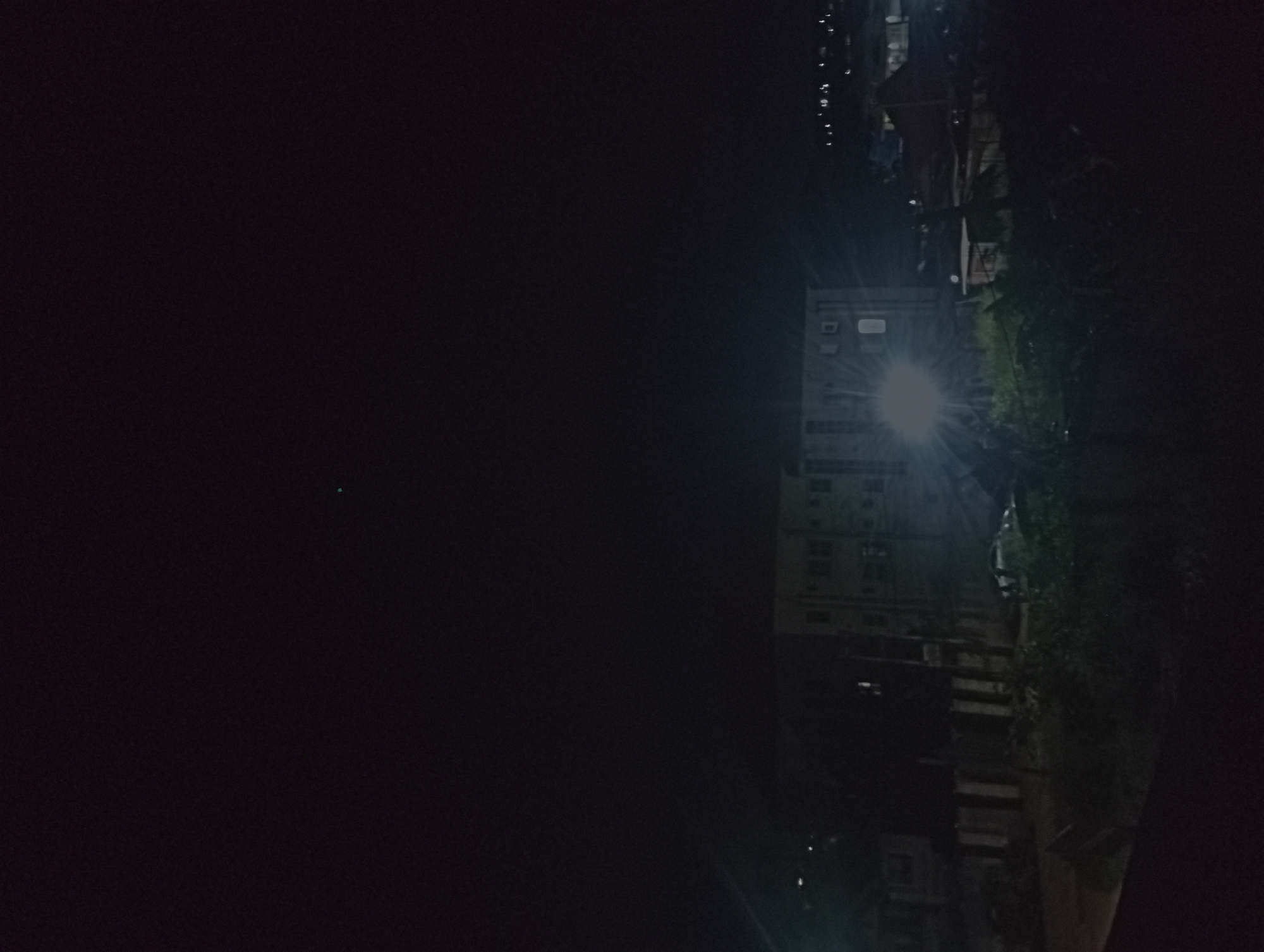 At night in Nkpor, Nigeria, a tall building stands next to trees or overgrown vegetation. A light source is visible on the building. The surrounding area is dark. Other buildings and objects are present in the distance, partially obscured by darkness.