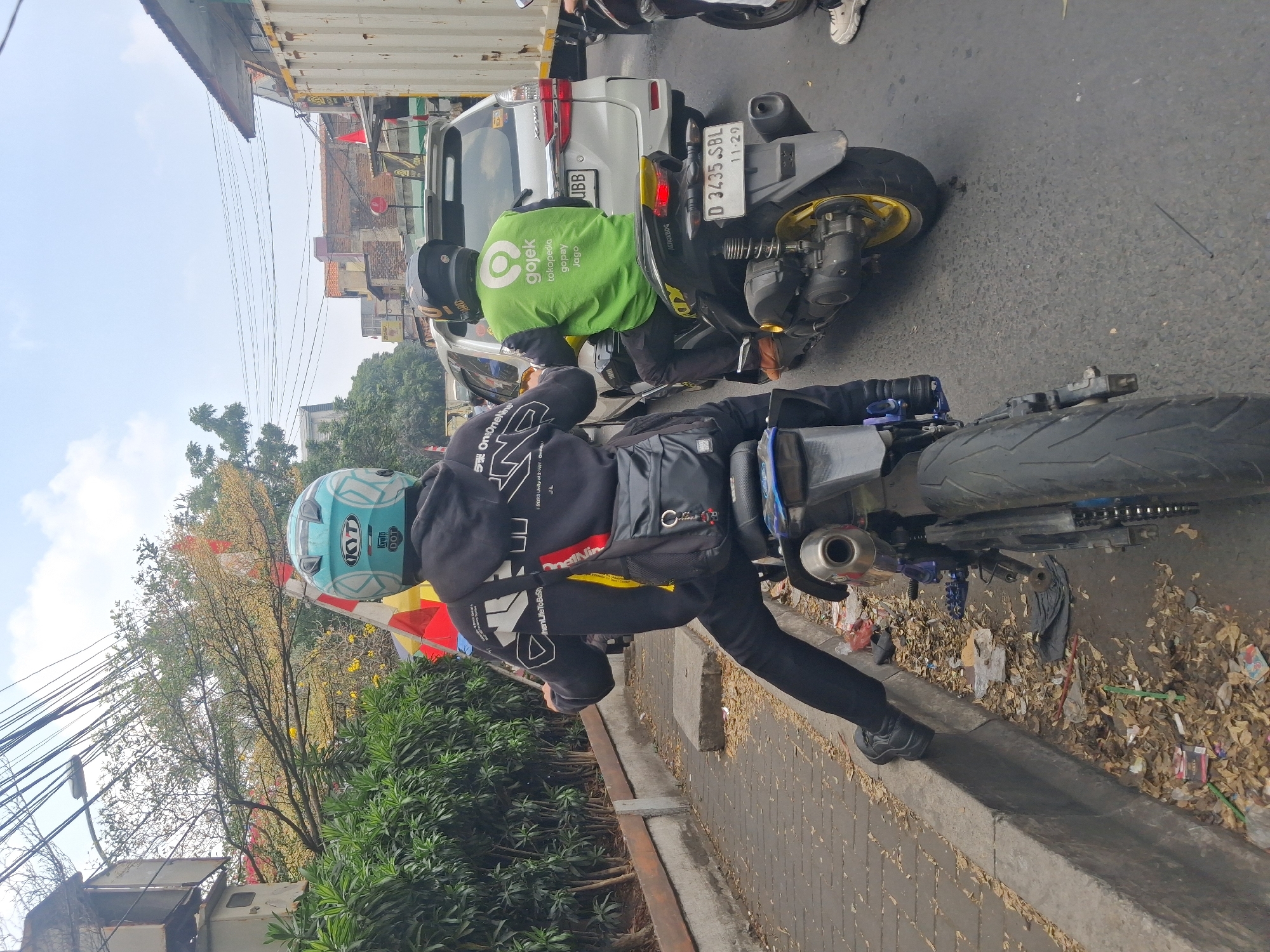 The image depicts a street scene in the city of Cimahi, Indonesia, likely during daylight hours given the sky's illumination. Two individuals on motorcycles are stationary in traffic. The motorcycle in the foreground is black with yellow accents, and the rider, wearing black pants, black shoes, and a black hoodie with white graphic prints, is partially visible from the rear. A black messenger bag is slung over their shoulder. They are wearing a light blue helmet with white decals, including the "KYT" and "DOT" logos.
The motorcycle in the middle ground is also black and yellow. The rider is wearing a bright green vest with the "Gojek" logo prominently displayed, indicating they are a delivery driver. They are wearing black pants and a black helmet. Behind them, a white SUV is also stopped in traffic.
The environment includes a paved road with visible traffic lines, a concrete sidewalk with some debris and fallen leaves, and lush green foliage lining the side of the road. Overhead, a complex network of electrical wires crisscrosses the sky, a common sight in urban Indonesian settings. Buildings and structures, including what appears to be a shipping container, are visible in the background, suggesting a commercial or mixed-use urban area. The overall atmosphere suggests a busy, potentially congested urban environment.