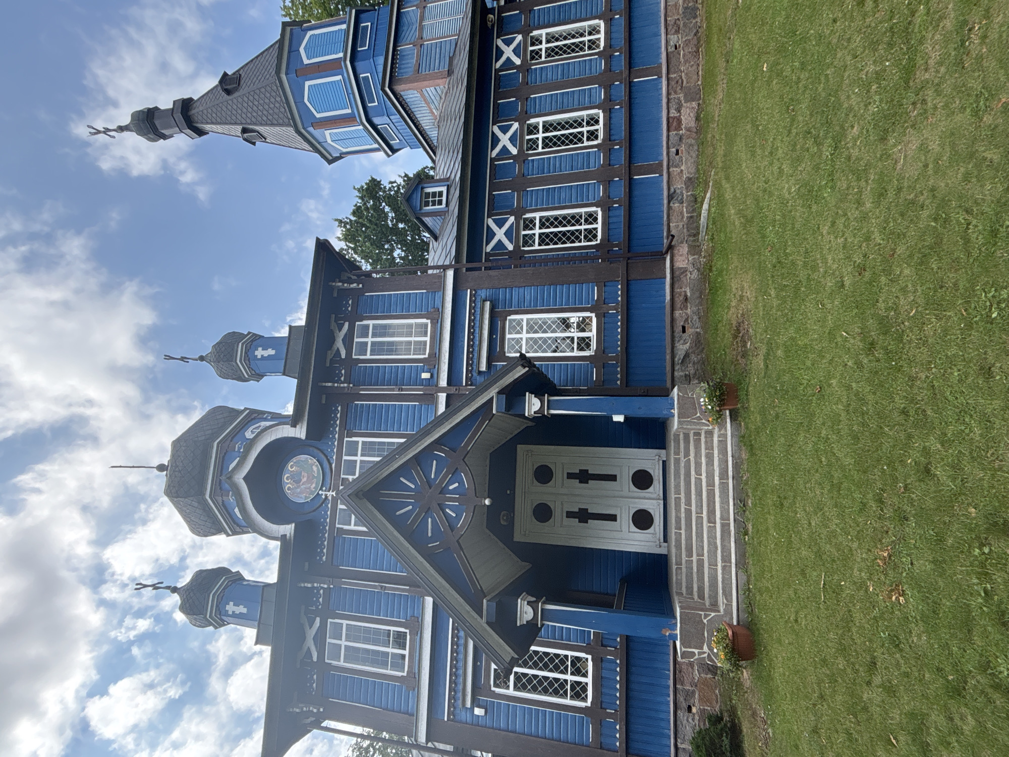 The image shows a bright blue wooden church with dark brown trim, located in Puchły, Poland. The church features multiple domes topped with crosses, and a prominent bell tower to the right. The facade of the church is adorned with decorative wooden carvings and features large, white-framed windows. A covered entrance with double doors, also painted white, is approached by a set of stone steps. Two potted plants with yellow and white flowers sit on either side of the steps. The church is surrounded by a green lawn, and the sky above is partly cloudy. The perspective of the photo is from a low angle, looking up at the structure.