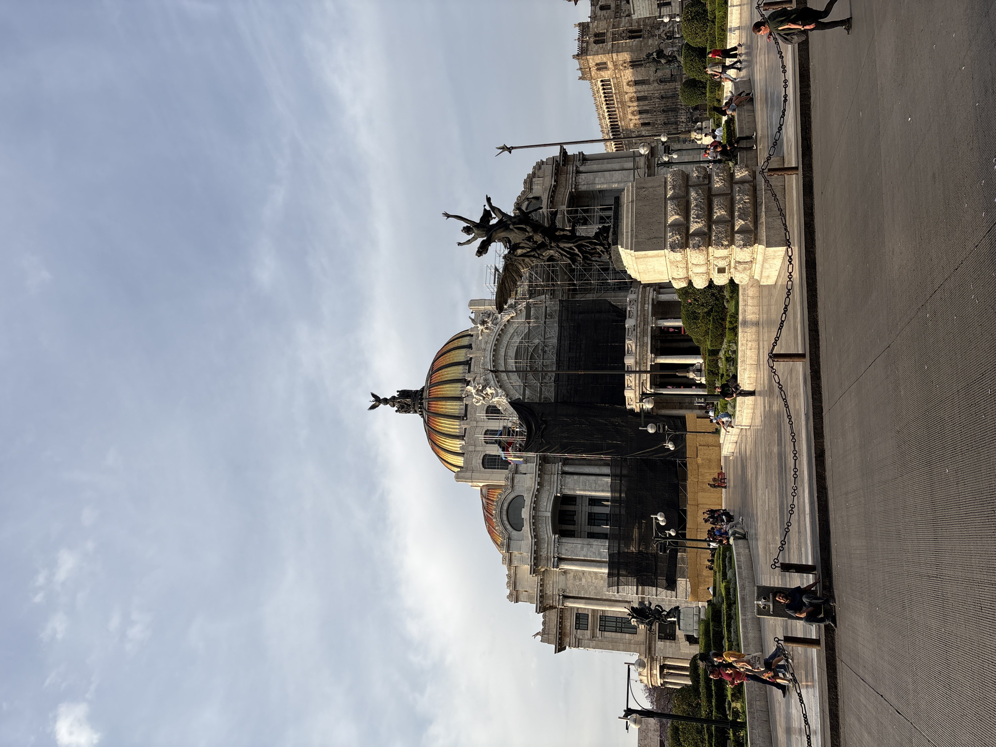 The image shows the Palacio de Bellas Artes (Palace of Fine Arts) in Mexico City, Mexico. The building is captured from a low-angle perspective. Scaffolding and black material cover a section of the building, presumably for maintenance or restoration.
In the foreground, there is a paved walkway, and a chain serves as a barrier. People are walking and sitting near the edge of the walkway. Further in the background, another building is partially visible. The sky is light blue with some clouds.