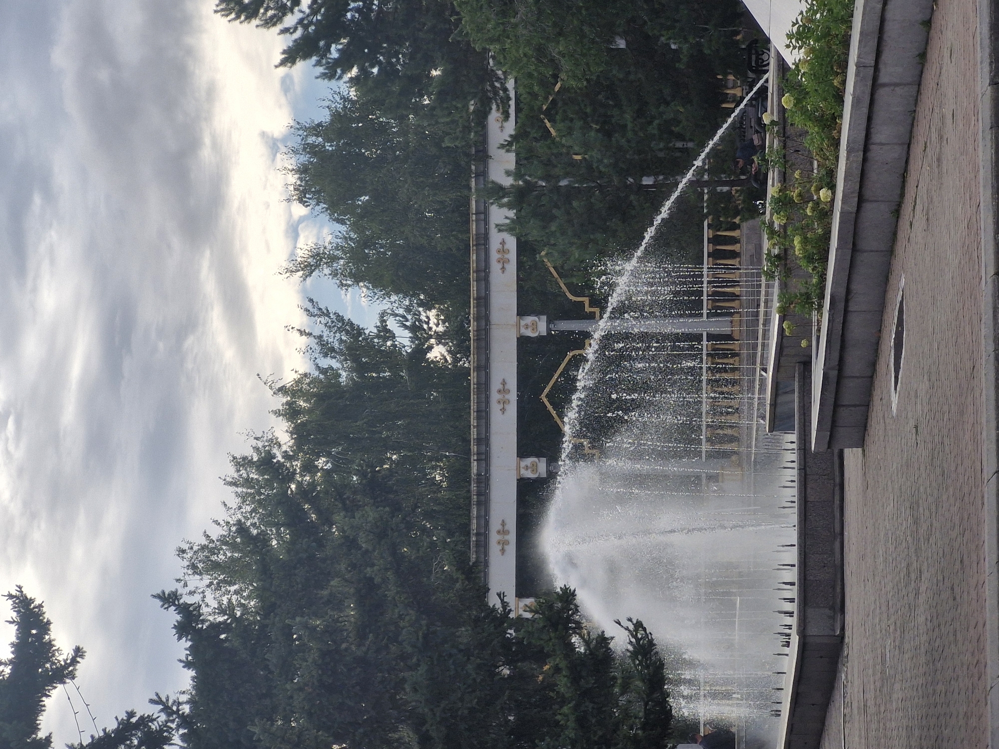 In Kokshetau, Kazakhstan, a fountain sprays water in an outdoor public area. The fountain has multiple jets, creating a curtain of water. The area around the fountain is paved with brick and bordered by concrete planters with green vegetation and flowers. In the background, trees and a decorative white and gold archway are visible. The sky is overcast with gray clouds. Two people are visible in the distance, one near the fountain and another sitting on a bench.