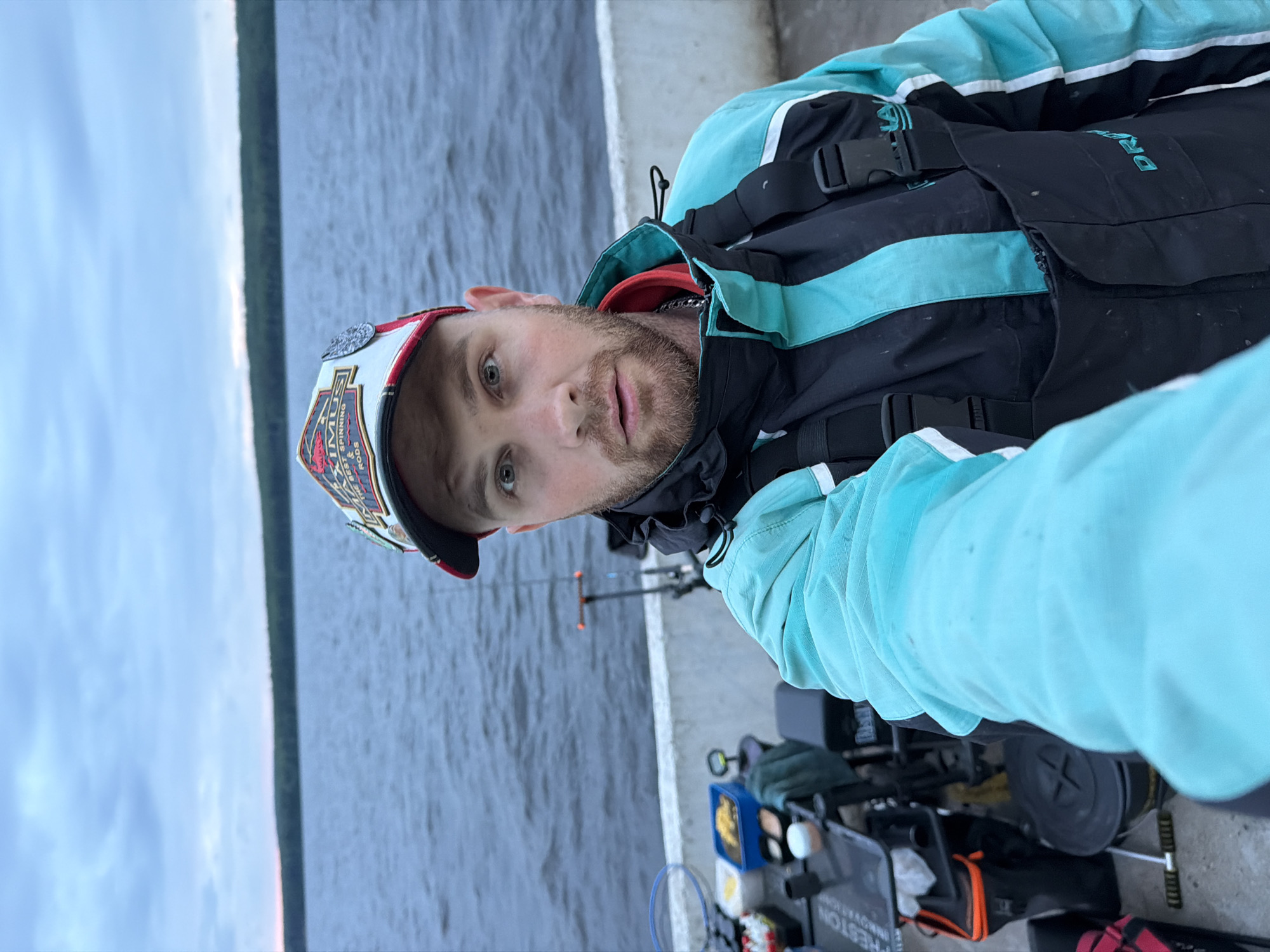The image captures a male individual taking a selfie outdoors in the city of Chaikovsky, Russia. The man is positioned centrally, captured from the waist up, extending his right arm to hold the camera. He has a light-brown beard and mustache, light-colored eyes, and an open-mouthed expression. He wears a multi-panel baseball cap with a red brim, featuring a white front panel emblazoned with a "MAXIMUS BEST SPINNING RODS" logo. His attire consists of a teal/aqua and black waterproof jacket over black fishing overalls with teal vertical accents and white stripes on the sides; black buckles are visible on the overall straps, and a metallic chain is around his neck.
To the left and behind the man, resting on a light grey concrete barrier that separates the viewer from a large body of water, is an array of fishing equipment. This includes a black equipment trolley or table displaying several containers (some white, some blue with yellow contents), a blue-rimmed fishing net, multiple fishing rods (one visible standing upright with an orange tip), and a black bucket. The branding "PRESTON INNOVATIONS" is discernible on some of the gear.
The background consists of a wide expanse of dark blue-grey water with ripples, stretching to a distant horizon. A line of dark green trees marks the far shoreline. The sky above is overcast, presenting a light blue-grey coloration with visible cloud patterns and a faint pinkish tint near the horizon, suggesting either dawn or dusk.