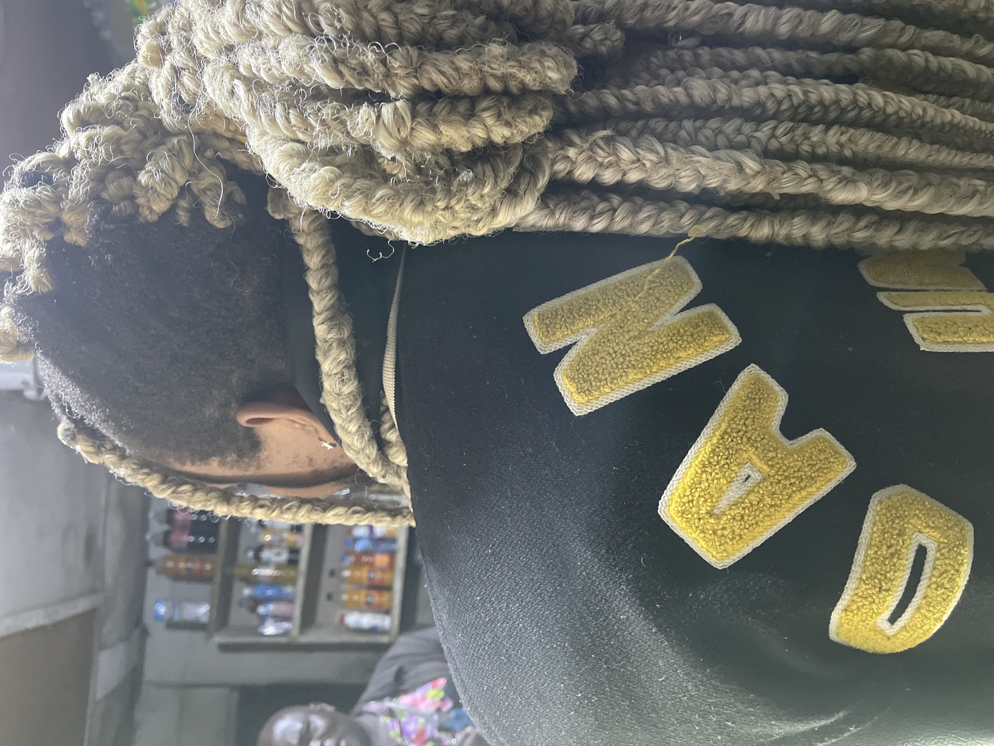 The image is a close-up, rear-view shot of a person with long, blonde braids. The person is wearing a black jacket with yellow, embroidered letters on the back that appear to spell "GOD" and possibly "JOIN". The person's hair is styled in numerous, thick braids that hang down their back. In the background, shelves are visible with bottles of beverages. Another person, appearing male with dark skin and wearing a floral shirt, is partially visible on the left side of the frame, looking towards the foreground. The environment suggests a small shop or a market stall. The overall impression is a candid moment captured within an urban setting.