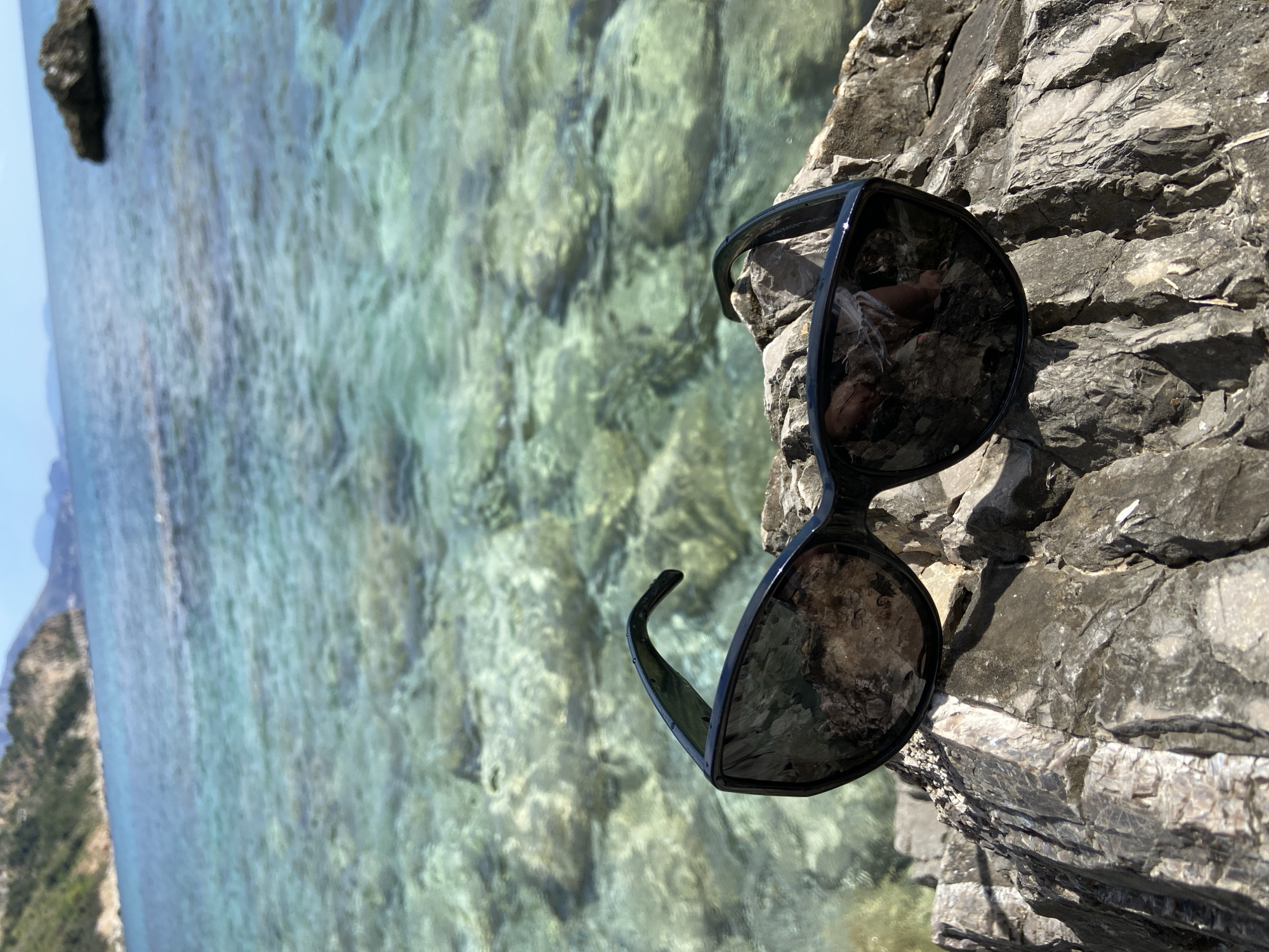 A pair of sunglasses rest on a gray, jagged rock face. The lenses reflect a blurred image of the rock. In the background, turquoise water stretches toward the horizon where a hazy coastline and distant hills can be seen. The location is most likely in the city of Prijevor, Montenegro.