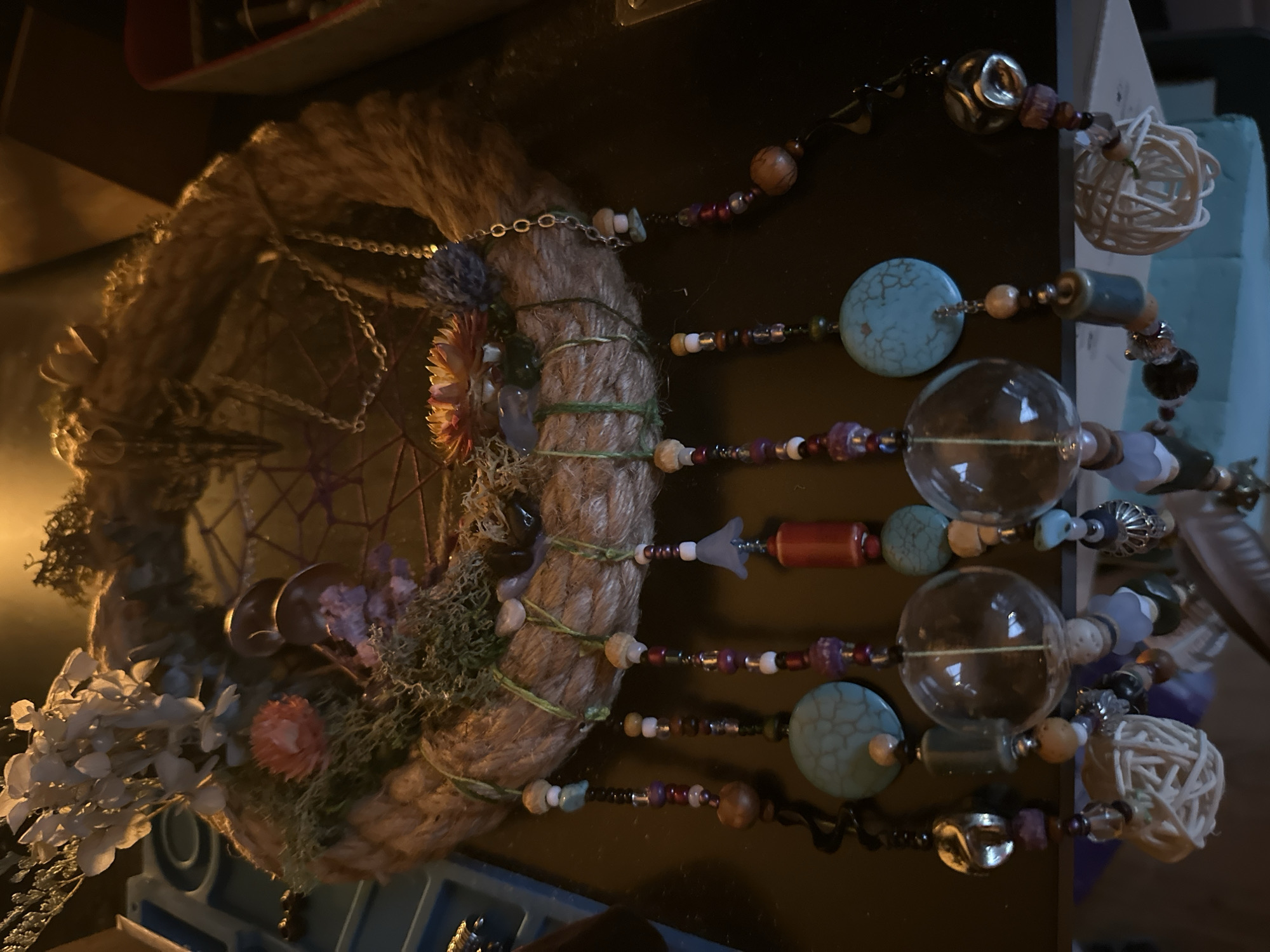 The image shows a close-up, overhead view of a handmade dreamcatcher-like object, likely a wind chime or wall decoration. The object is circular and appears to be constructed from thick, braided rope. It is adorned with various natural elements such as dried flowers, moss, and what look like small pinecones or seed pods. Dangling from the bottom of the circular frame are several strands, each made of numerous beads of different shapes, sizes, and colors, including turquoise, wood, glass, and metallic elements. Some strands also feature larger decorative elements like clear glass orbs, a cylindrical red bead, and intricate woven natural fiber spheres. The background is dark and appears to be a tabletop or shelf. The lighting is warm and casts shadows. There is no discernible location within McMasterville, Canada evident in the image.