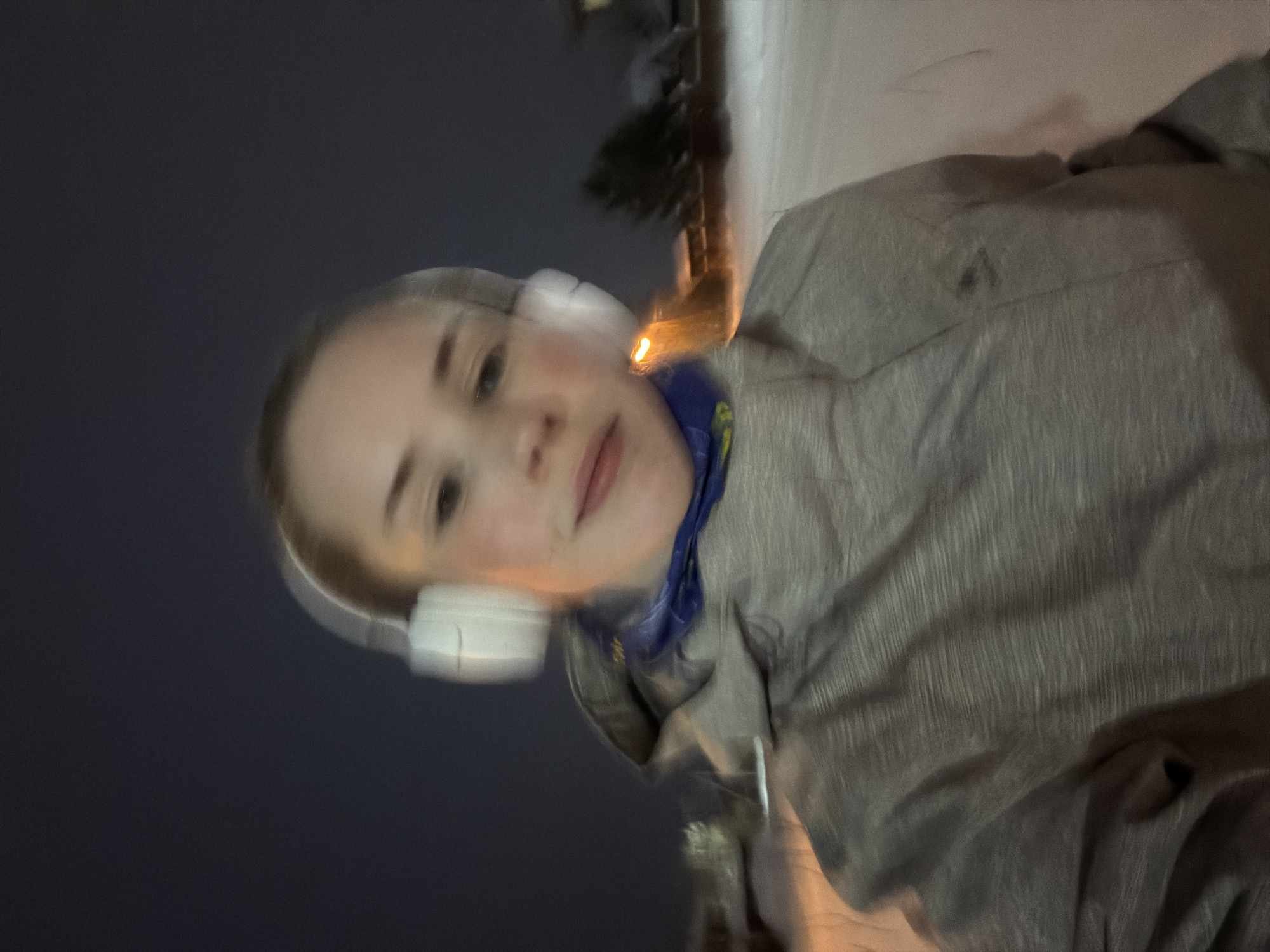 A blurred, selfie-style photograph shows a fair-skinned person with headphones, wearing a grey hooded jacket and blue neck gaiter. The background is dark, transitioning to a snow-covered ground with a dark tree in the upper center. The photo may be taken in Berezniki, Russia at night or during low light conditions. No specific actions or interactions are discernible due to the blur.