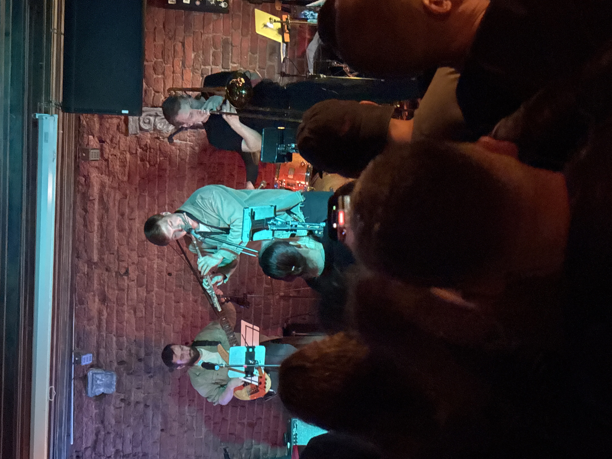 The image depicts a band performing on a stage in front of a brick wall. The lighting is dim, with blue and pink hues illuminating the scene. In the foreground, a crowd of people is visible from behind, with their backs turned to the camera. The audience appears engaged with the performance.
On stage, three musicians are actively playing. The musician on the left is playing a bass guitar and is wearing a light green jacket. The musician in the center is playing a flute and is also dressed in a light green jacket. The musician on the right is playing a trombone and is wearing a black shirt. A drum set is partially visible behind the central musician. Electronic equipment, including speakers and monitors, is also present on the stage. A laptop is on a stand to the right of the trombone player.
The venue appears to be a live music club or bar. The brick wall suggests an industrial or vintage aesthetic. The presence of a stage, instruments, and an audience indicates a concert or live performance setting.
While the image itself doesn't provide explicit geographical markers, if this scene were to be located in Minsk, Belarus, it would likely be in a venue known for hosting live music. Such venues in Minsk could range from dedicated concert halls and clubs to bars and restaurants that feature live entertainment. For instance, a place like "Re:Public" or "Bruges" in Minsk are known for hosting concerts and could potentially fit this description, although without further context, pinpointing the exact location is not possible. The atmosphere suggested by the image is one of a lively, intimate musical performance.