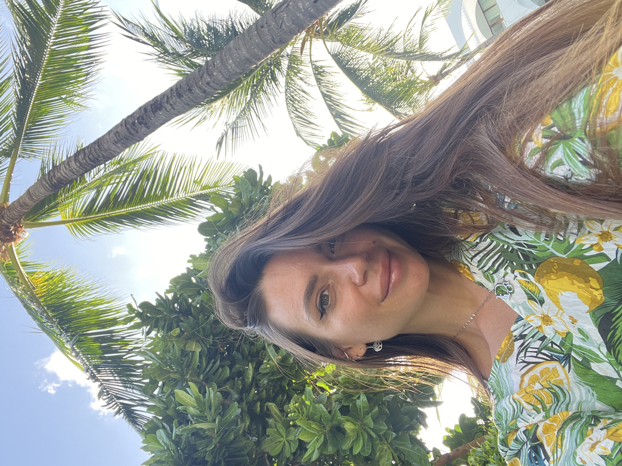 An outdoor, low-angle, medium shot shows a woman with long brown hair pulled to the right, her face turned to the left, in front of lush green foliage and palm trees against a bright sky. She wears a tropical-print garment with yellow and green lemons and white flowers, a silver necklace with a 'D' charm, and small earrings. The woman has on eyeliner and lipstick. A building is visible in the background.