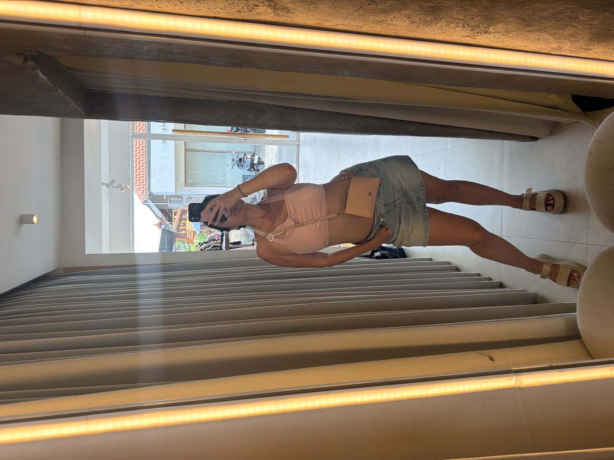 A woman is taking a mirror selfie in a brightly lit room, possibly a changing room or a boutique, in North Kuta, Indonesia. She is wearing a pink halterneck crop top, a light blue denim mini skirt, and beige platform sandals. A small beige crossbody bag is slung over her shoulder. The room features cream-colored vertical blinds and a concrete accent wall with a vertical fluorescent light. Through the doorway in the background, a street scene is visible with buildings and a scooter.