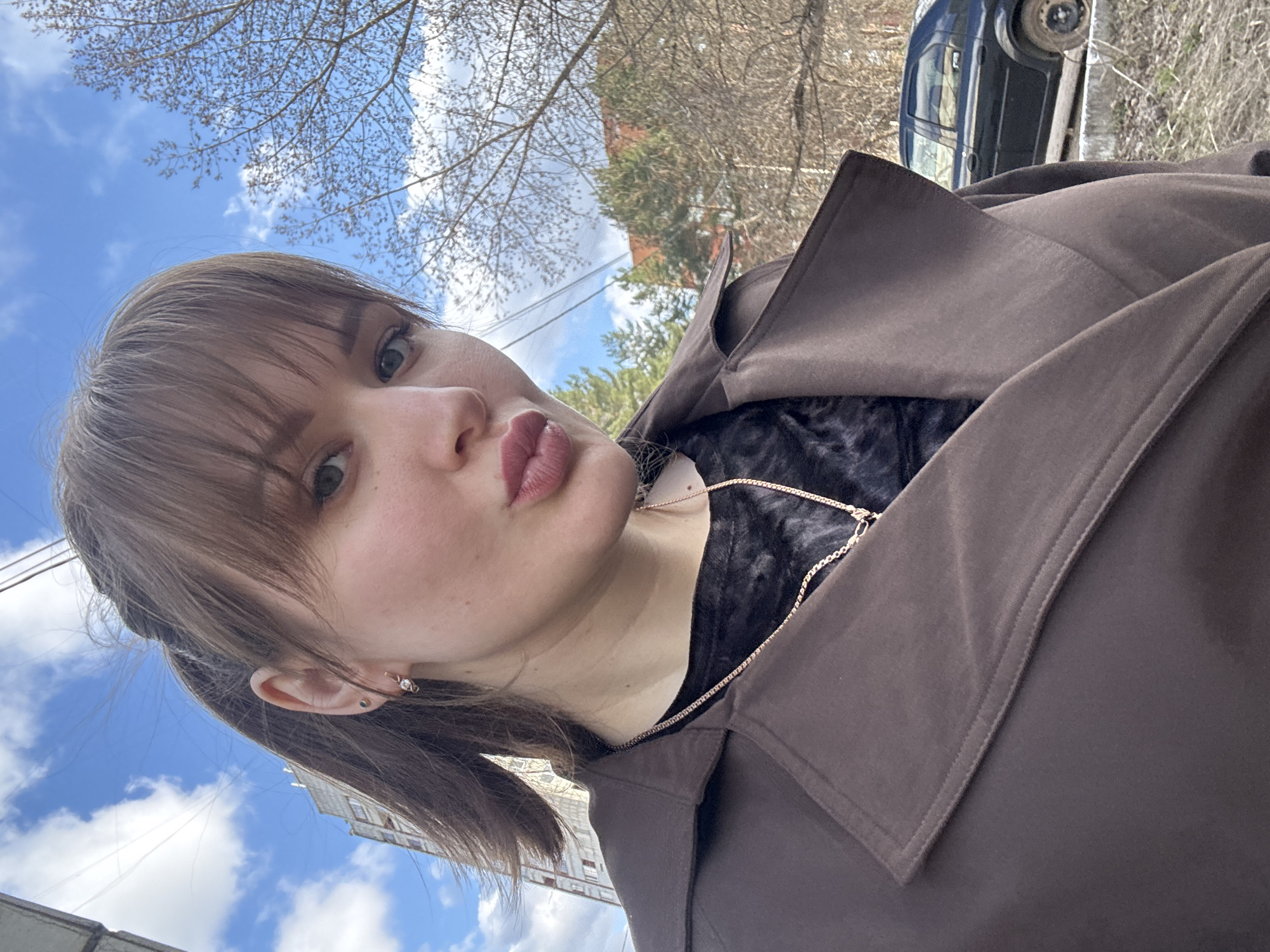 An outdoor selfie captures a young woman from a low-angle perspective, with the image rotated 90 degrees clockwise, making the sky appear to her right. The woman, with light brown hair and bangs, is looking directly at the camera with a playful expression, her lips puckered into a "duck face." She has light-colored eyes and small earrings, one appearing to be a hoop and another a stud. She is wearing a dark brown, possibly trench-style, coat with a large collar, over a dark, subtly patterned top. A delicate gold-colored chain necklace is visible around her neck.
The background features a clear blue sky with fluffy white clouds, indicating fair weather during the daytime. Bare tree branches, typical of early spring or late autumn, are prominent against the sky and extend across the upper part of the frame (when viewed correctly). In the mid-ground, a multi-story building, possibly an apartment block, is visible, and further to the right, a dark-colored car is parked on what appears to be a dirt or grassy area next to a road. The scene suggests an urban or suburban setting, possibly a street in Chaikovsky, Russia, as provided by the context. No visible text is present in the image.