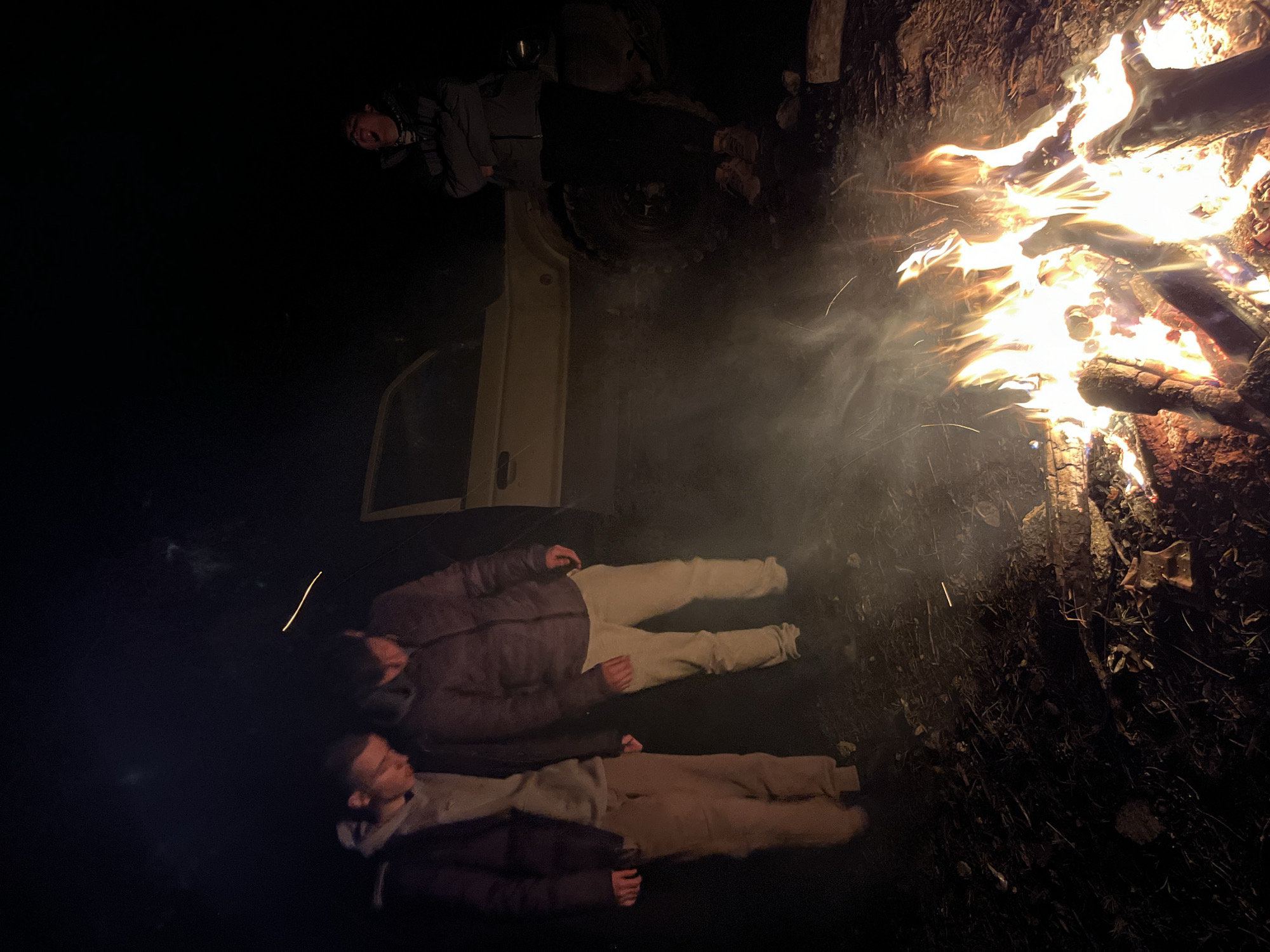 Two people are lying on the ground beside a campfire. A vehicle door is open nearby, and another person stands near the vehicle. The location is obscured, but inferred to be Karavelovo, Bulgaria.