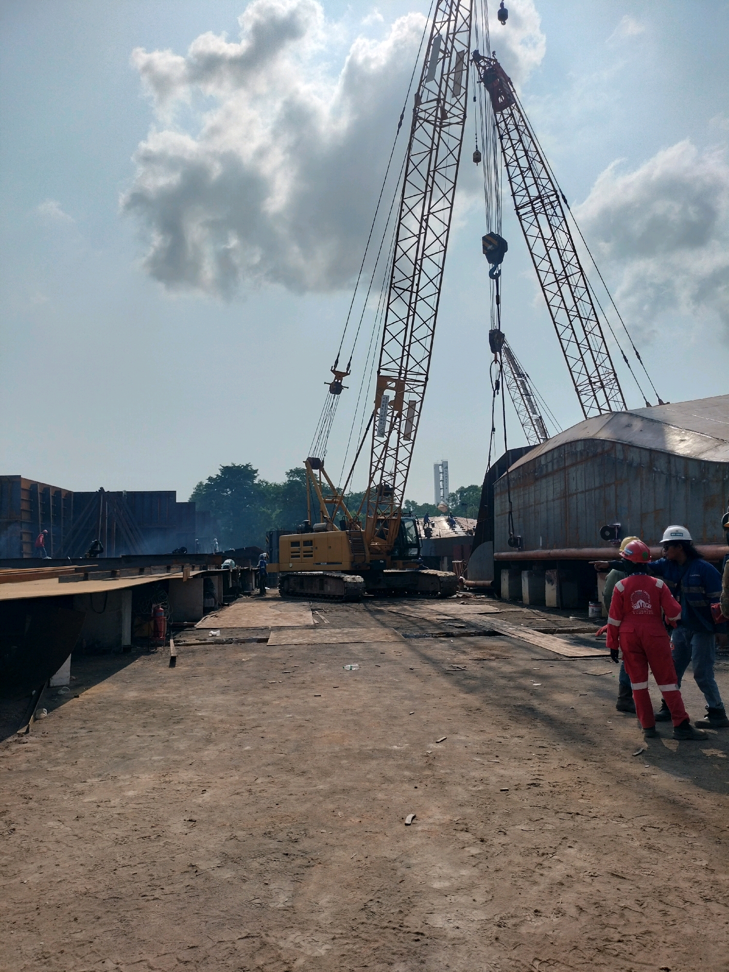 The image depicts an outdoor industrial construction or repair yard, likely within the city of Batam, Indonesia, given the context of shipbuilding and heavy industry prevalent in the region. The scene is dominated by large construction equipment, specifically two massive cranes with towering lattice booms that converge towards the sky. A yellow crawler crane with its boom raised is visible in the mid-ground, positioned on a dirt and gravel surface. In the background, large metal structures, possibly ship hulls or modular components, are under construction or assembly, with some sections appearing to be steel plating.
Several individuals are present, all wearing personal protective equipment, indicating they are workers. In the foreground, three individuals are clustered together, two of whom are engaged in conversation. One worker, wearing a bright red jumpsuit, is gesturing with their hand. Another worker, in a blue uniform, stands beside them. A third individual, partially obscured, is also in the vicinity. Further into the yard, other workers can be seen scattered around the construction site, some appearing to be performing tasks or supervising.
The environment is characterized by a dusty, unpaved ground surface, common in such industrial settings. There are scattered pieces of wood and metal debris. The sky is partly cloudy, with bright sunlight casting shadows. A tall, slender tower structure, possibly part of a distant industrial facility or a communication tower, is visible on the horizon in the background, consistent with an industrial landscape. The overall impression is one of ongoing heavy industrial activity in a shipyard or a similar large-scale fabrication area.