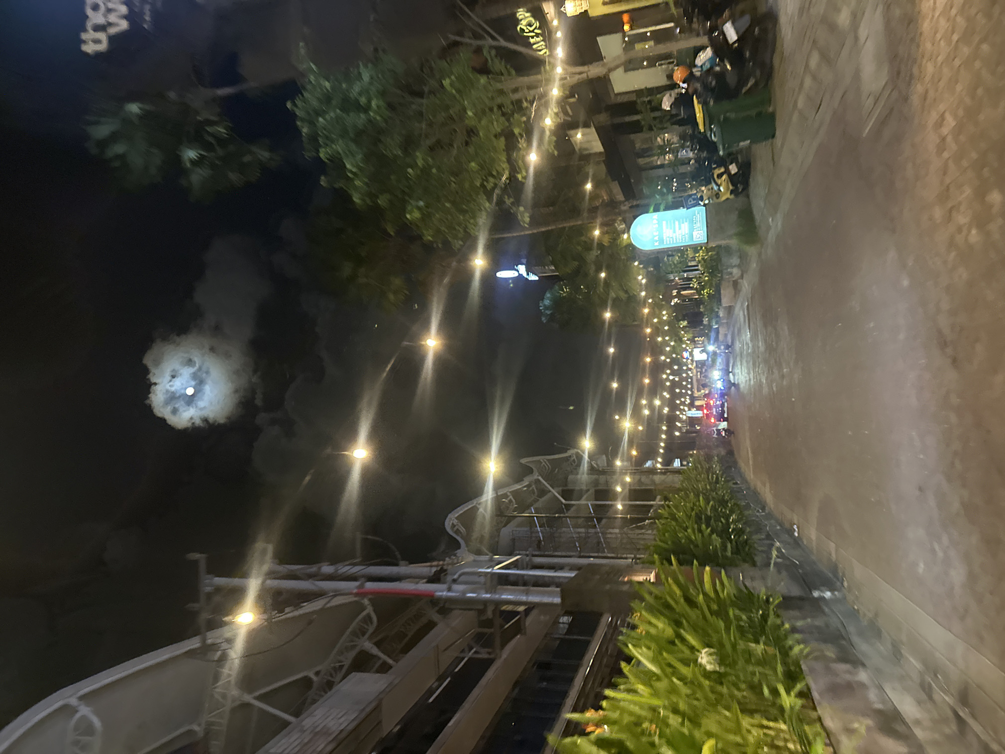 A nighttime street scene in North Kuta, Indonesia, shows a road lined with buildings and lush green foliage. String lights are draped across the street, casting a warm glow on the scene. The moon is visible in the night sky, peeking through the clouds. Several motorcycles are parked along the side of the road, suggesting this is a popular area for locals and tourists. One motorcycle has an orange helmet on its seat, and another rider is partially visible in the background. A bright blue sign with text that reads "KAE SPA" is mounted on a pillar. The buildings appear to be shops or restaurants, with some displaying signs and architectural details that suggest commercial activity. The overall atmosphere is lively and inviting, with the lights creating a festive ambiance.