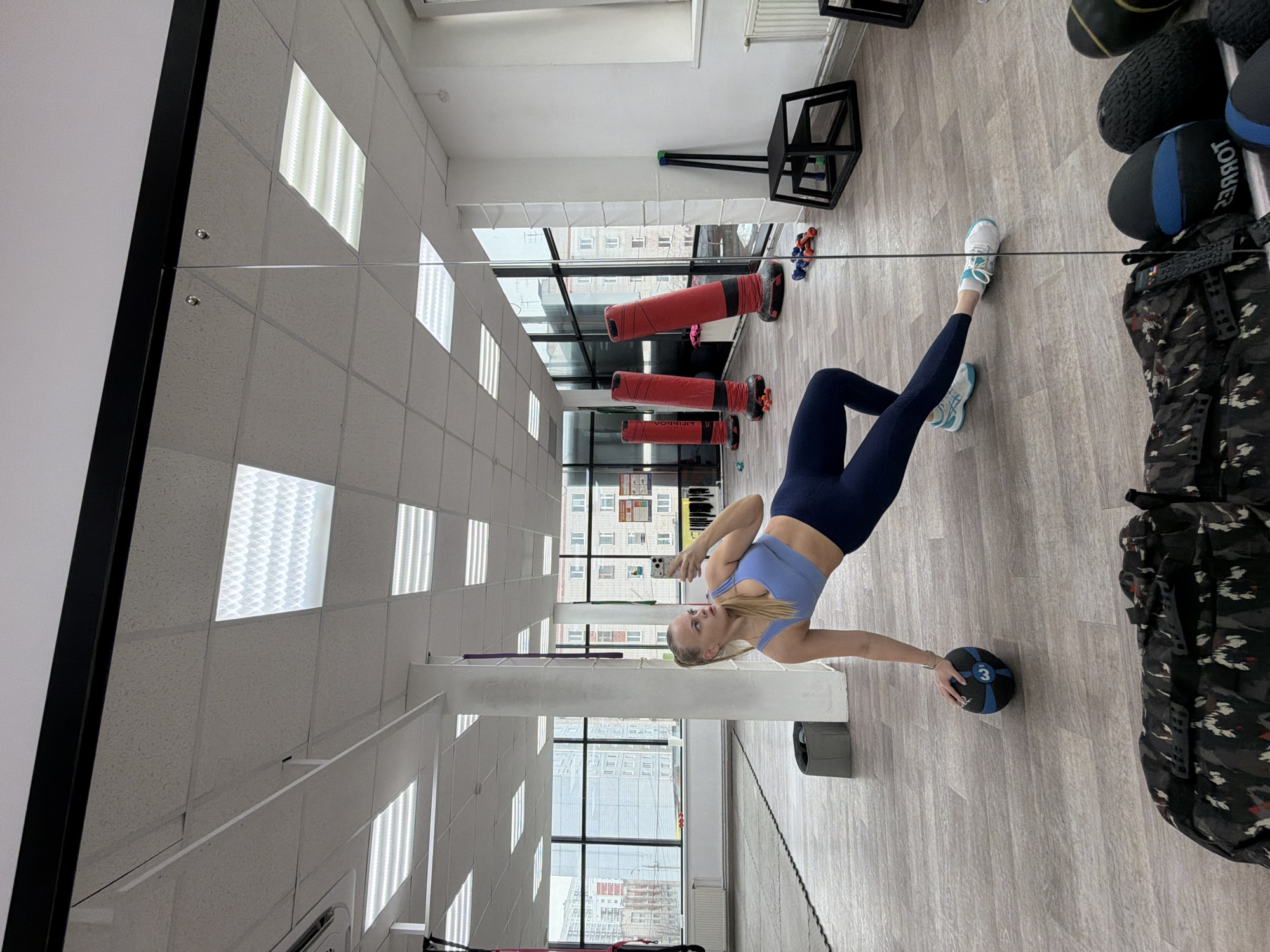 A woman is in a gym in Izhevsk, Russia. The woman has blonde hair and is wearing a light blue sports bra and dark blue leggings. She is holding a black and blue medicine ball with the number "3" on it. The gym has fluorescent lighting and a gray laminate floor. There are red punching bags, a black plyometric box, and medicine balls. A camo backpack is also visible. Buildings outside the gym windows indicate this is Izhevsk, Russia.