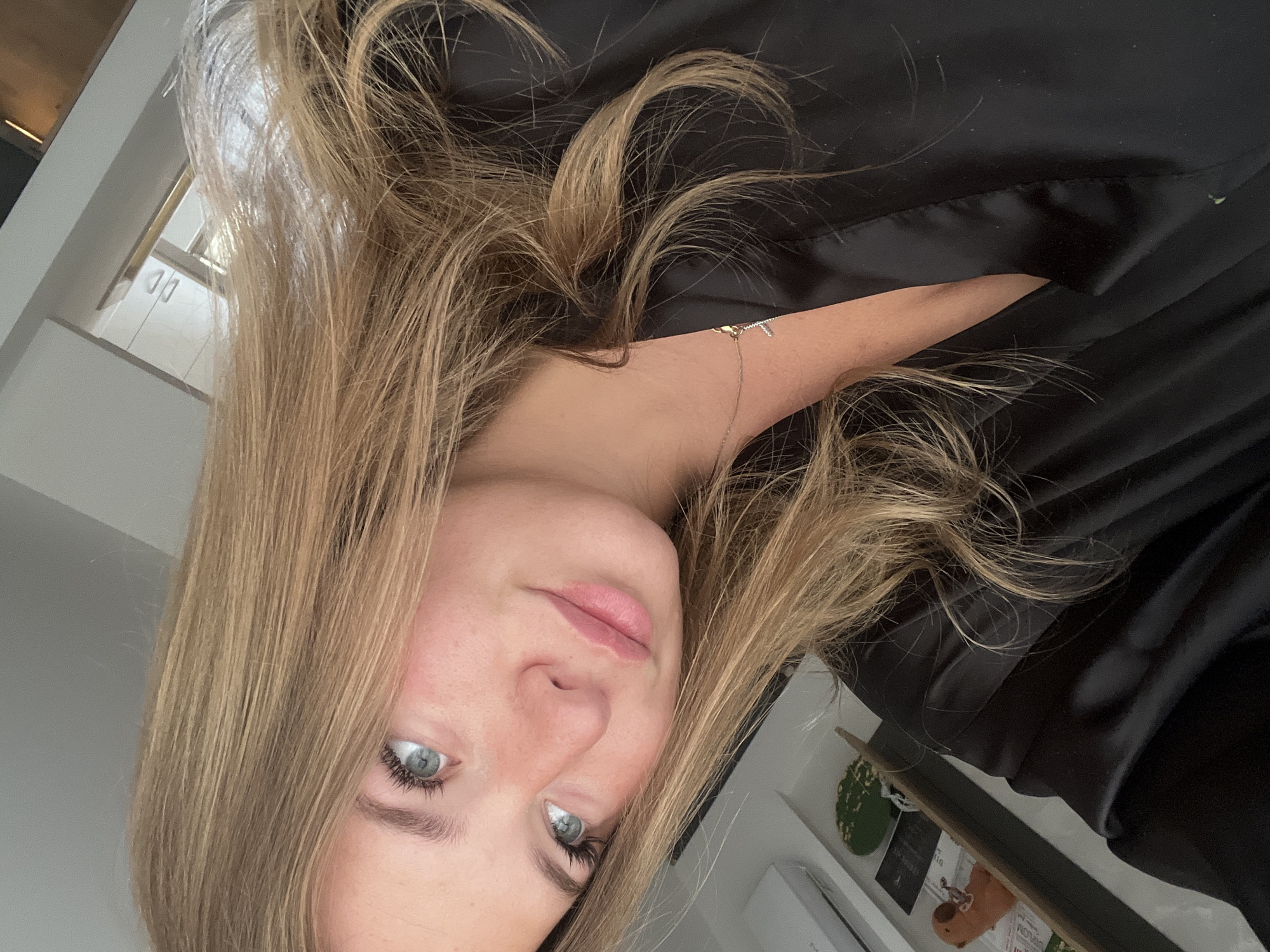 The media file is a close-up selfie of a woman with long, wavy blonde hair and striking blue-grey eyes. Her head is tilted slightly, and she looks directly at the camera with a relaxed, natural expression. She has clear skin and her lips are adorned with light-colored lip gloss.
She is wearing a black, possibly satin or silk, garment that offers a sharp contrast to her light hair and complexion. A delicate gold-colored necklace with a small pendant, possibly a letter or a cross, rests around her neck.
The setting appears to be indoors, likely a home, with soft, natural light illuminating her from the left. The background features light-colored walls or surfaces. To the right, a glimpse of a shelf or surface shows a small green potted plant and other indistinct objects. In the upper left, architectural elements, possibly window frames, are visible. The image conveys a casual and intimate atmosphere. No discernible text is visible within the frame.