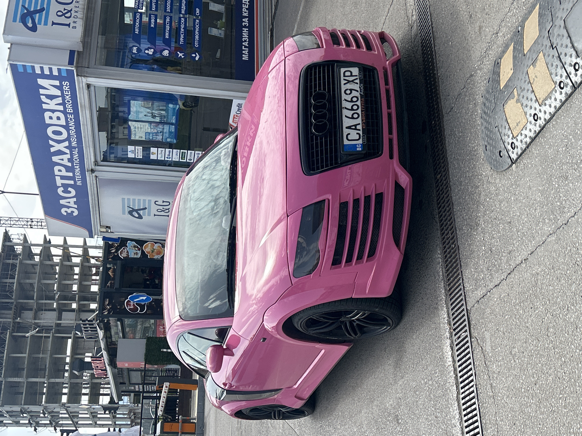 The image features a pink Audi TT car parked on a street in an urban environment, likely in Unknown, Unknown. The car is positioned on the right side of the frame, facing towards the top of the image. It has a Bulgarian license plate with the number "CA 6669 XP". The front of the car is heavily customized with a custom front bumper and blacked-out grill.
Behind the car is a building with a sign that reads "I&C BROKERS INTERNATIONAL INSURANCE BROKERS" in blue and white. The signage also lists various types of insurance in Bulgarian. The building also has other storefronts visible.
In the background, there's a tall, unfinished building structure made of steel beams.
The street surface is made of asphalt and has a drainage grate along the edge. A speed bump is partially visible on the upper right corner of the image.