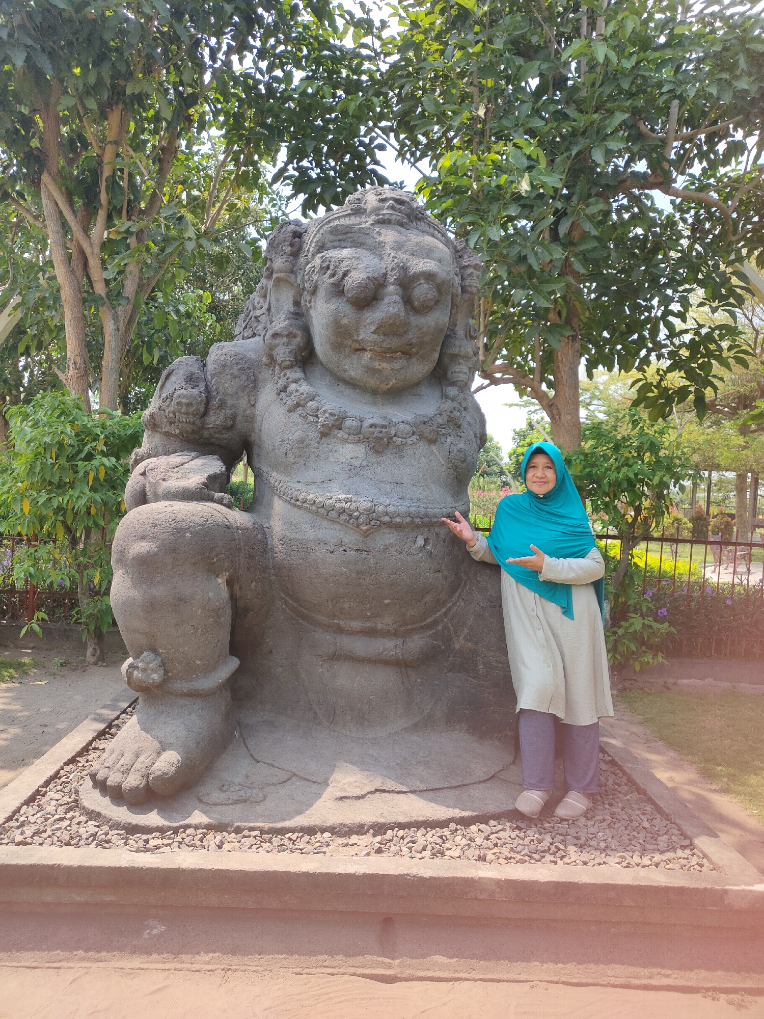 A woman wearing a light green tunic, dark pants, and a teal hijab stands next to a large, weathered stone statue in Pagu, Indonesia. The statue depicts a grotesque figure with bulging eyes, a wide mouth, and a necklace made of skulls. The woman is smiling and gesturing towards the statue with both hands. The statue is positioned on a gravel base within a fenced area, with trees and lush vegetation surrounding it. The ground is sandy, and the sky is a clear blue.