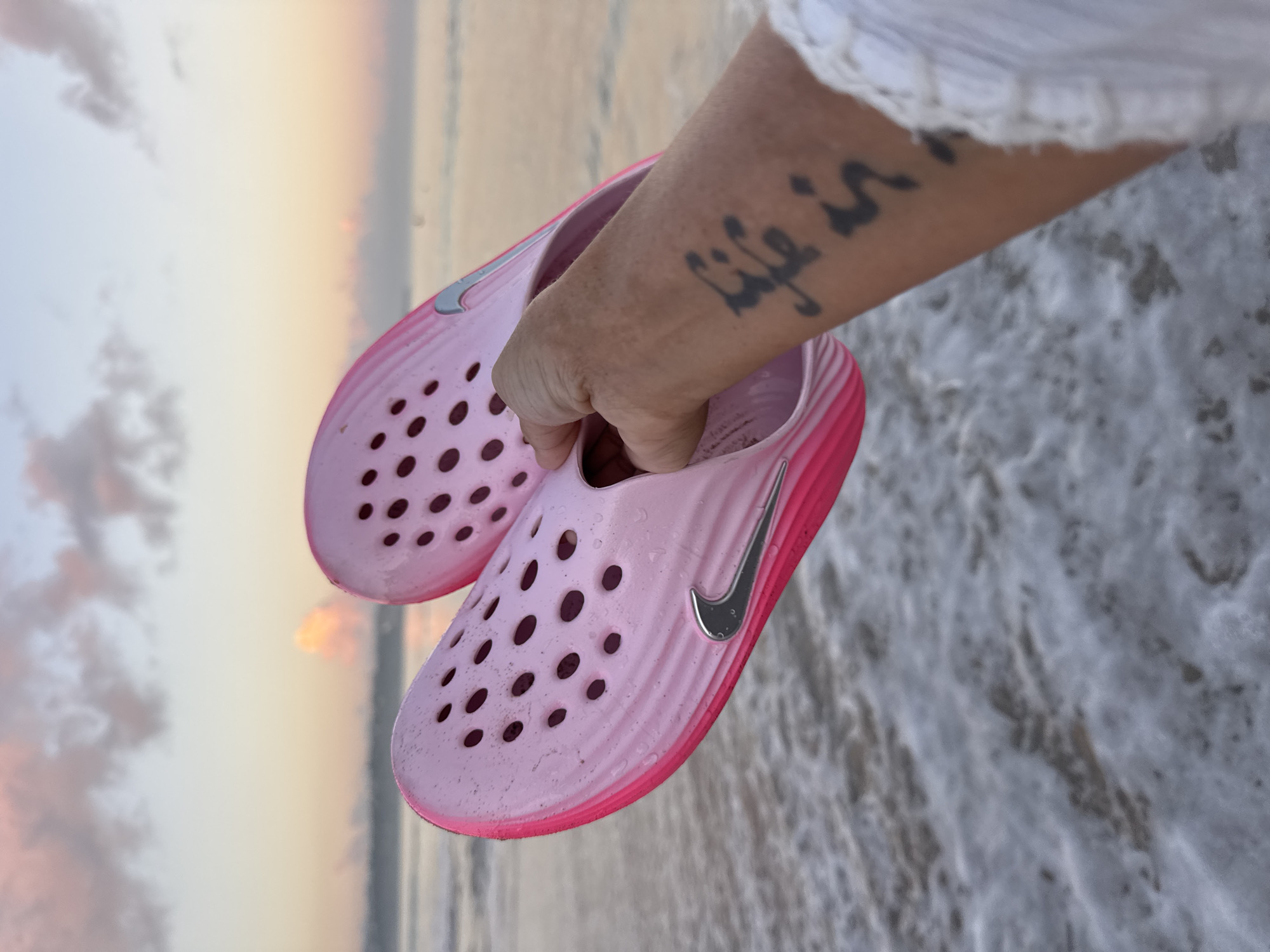 A close-up, angled shot captures a person's arm and hand holding two bright pink and silver Nike slip-on shoes over the ocean at what appears to be either sunrise or sunset in Kuta, Indonesia. The shoes feature numerous circular ventilation holes and prominent silver Nike swooshes on their sides. The hand is gently gripping one of the shoes.
On the person's forearm, a tattoo is visible, reading "Life is..." with the rest of the text partially obscured. A white, possibly lacy or textured, garment covers the person's shoulder and upper arm, visible at the top right of the frame.
The background reveals a tranquil beach scene. The ocean water below the shoes is frothy with white foam from gentle waves. The sky displays a soft gradient of pastel colors, transitioning from light blue and greyish clouds at the top left to warm oranges and pinks near the horizon, typical of dawn or dusk. The overall atmosphere is peaceful and serene, suggesting a moment of relaxation by the sea.