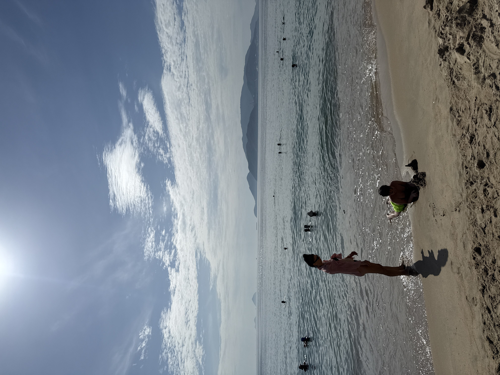 The image depicts a beach scene in Nha Trang, Vietnam, on a sunny day with a bright sun visible in the upper left corner and scattered clouds. The sea is calm with gentle waves lapping at the shore. Several people are visible in the water, swimming and enjoying the ocean. In the foreground, on the sandy beach, two individuals are present. One, a young person wearing a pink cover-up and sunglasses, stands near the water's edge, looking down. Beside them, a young person wearing green swim trunks lies on their stomach, playing with sand. The background features a coastline with lush, green mountains under a hazy sky. The overall atmosphere is one of a peaceful, recreational day at the beach.