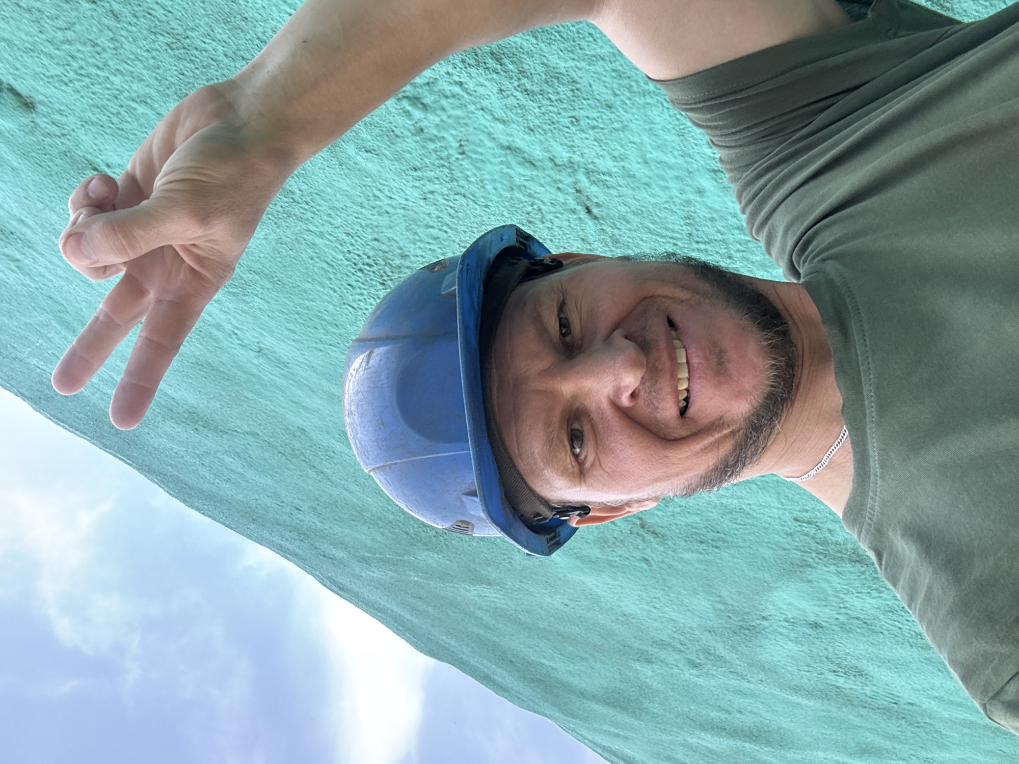 A man wearing a blue hard hat and a green t-shirt stands in front of a textured teal wall under a cloudy sky. He is smiling and holding up his hand in a peace sign. The man has short brown hair and a beard. He is also wearing a silver chain necklace. The image appears to have been taken outdoors, possibly in a construction area. There is no visual information to suggest the location is in Oskemen, Kazakhstan.