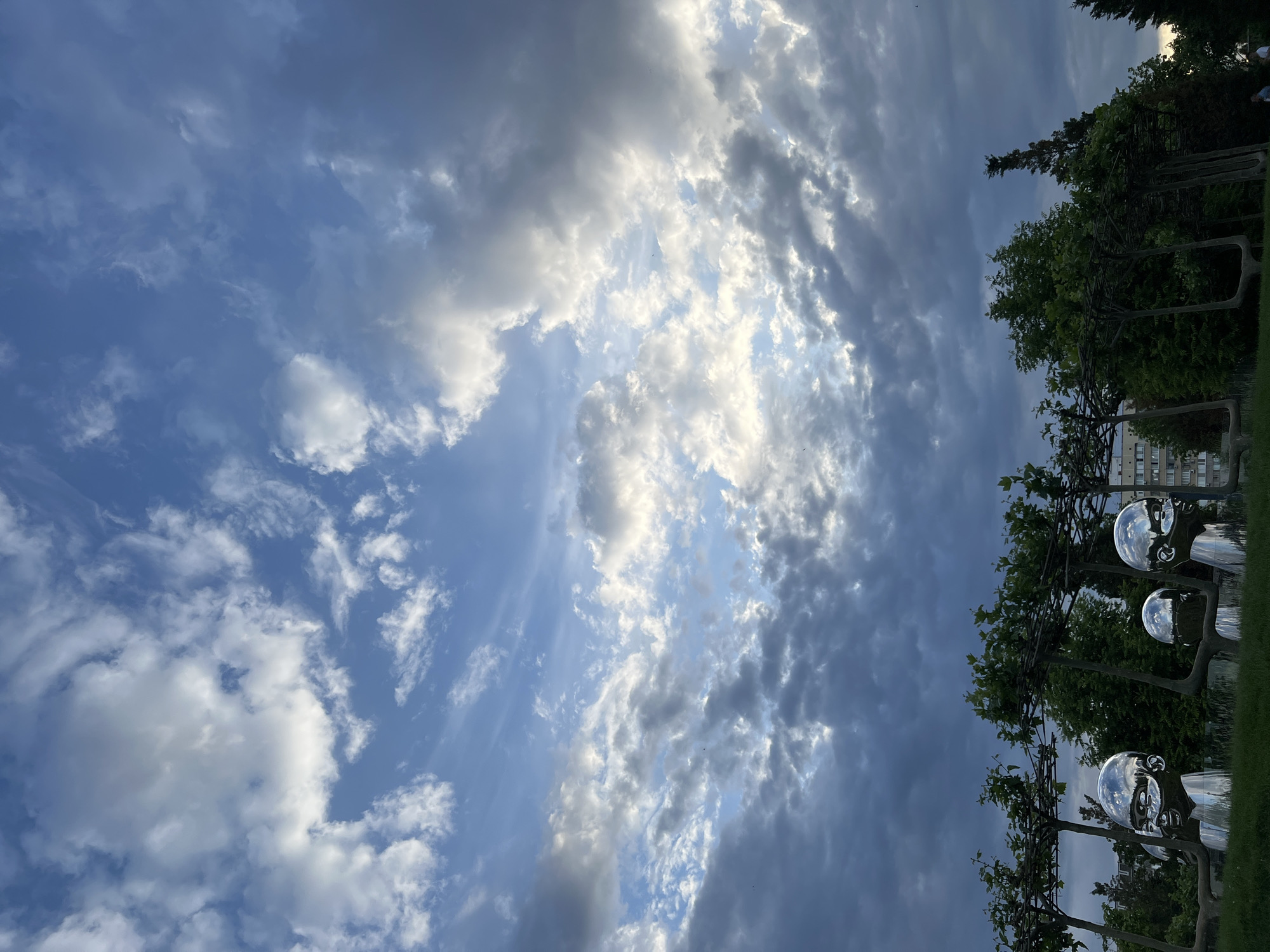 The image captures a vibrant, partially cloudy sky above a park landscape, likely during late afternoon or early evening. The sky is dominated by a dramatic interplay of bright white, fluffy cumulus clouds and darker, denser grey clouds. Patches of clear blue sky are visible, particularly towards the upper left. Sunlight breaks through the clouds, creating strong highlights and casting parts of the sky into shadow. Small, dark specks, possibly birds, are scattered across the sky.
In the lower portion of the image, there is a lush green landscape featuring several trees with dark green foliage. Some trees appear to be espaliered or trained on elaborate metal structures, giving them a distinct, architectural form. Among these trees, on the right side, are multiple large, spherical, reflective sculptures. These sculptures, resembling polished metallic heads or large eyes, mirror the surrounding sky and trees, creating interesting distorted reflections. A strip of green grass is visible at the very bottom. In the background, through the trees, a light-colored building with multiple windows is faintly discernible. There are also very faint figures of people in the distance, behind the trees.
The scene suggests a well-maintained public park or garden, characterized by both natural elements and artistic installations. Given the context, this is likely Sergey Galitsky Park in Krasnodar, Russia, known for its unique landscapes and art pieces. No visible text is present in the image.