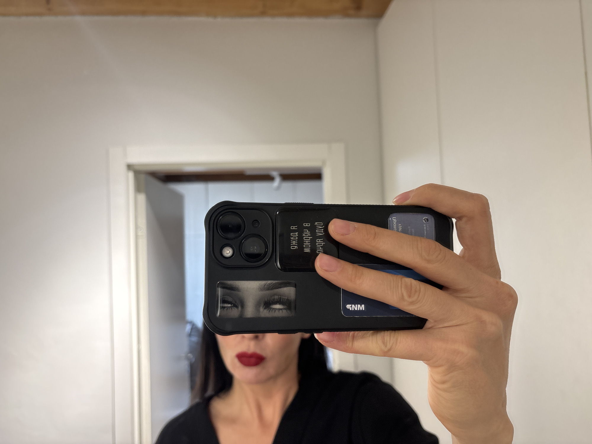 The image shows a woman taking a selfie in a bathroom or similarly furnished room. The phone, an iPhone in a black protective case, obscures the majority of the woman's face, but red lipstick is visible. The phone case has a picture of upward-gazing eyes on it, and the reverse side of the case has writing on it in a non-Latin script, along with the initials "GNM". A credit card or similar card is also visible on the back of the case. The room has white walls and a white door frame in the background, with a glimpse into an adjacent room visible through the doorway. The room has a partially visible wooden ceiling. There is no concrete information about the city of Chaikovsky.