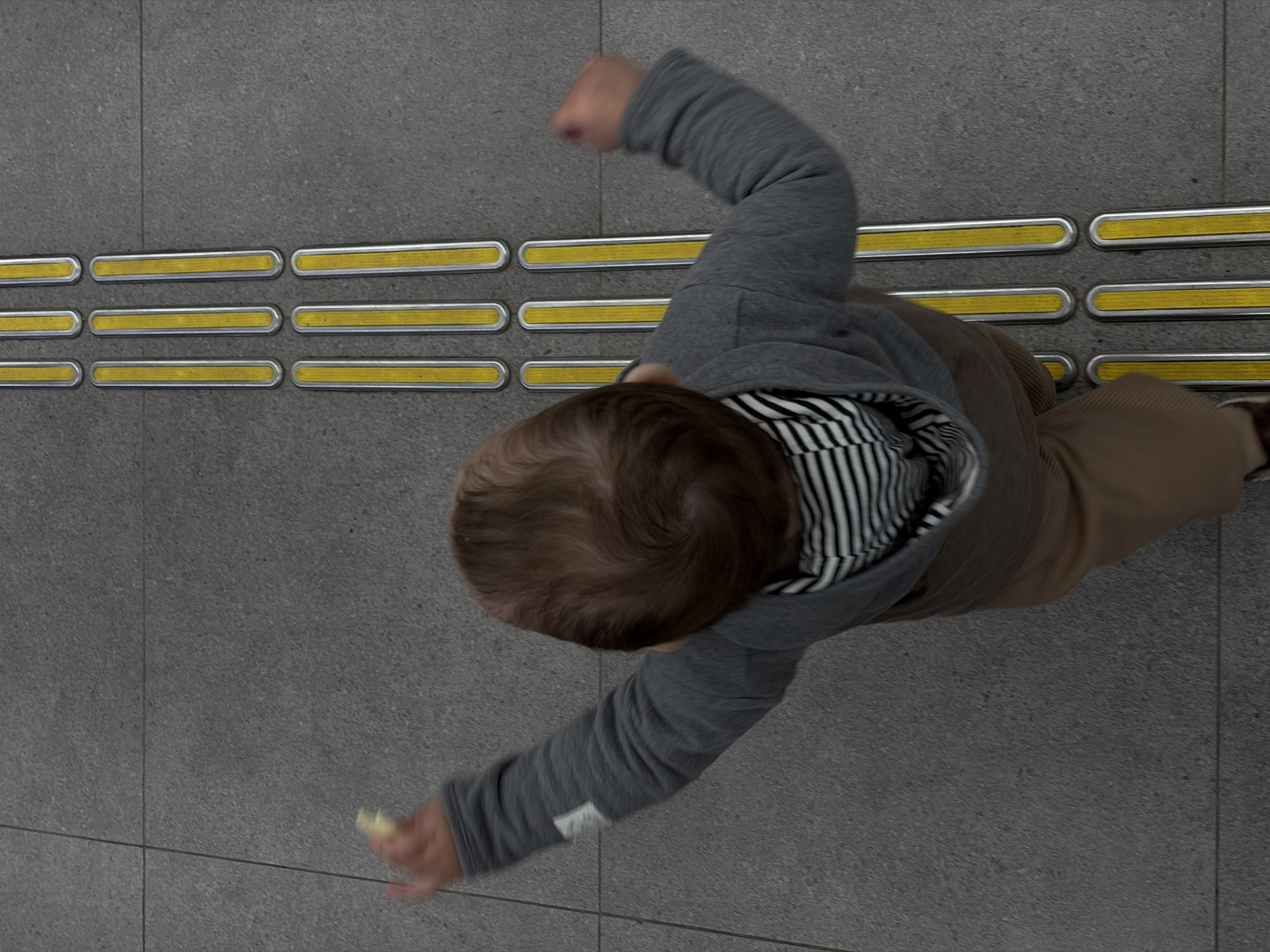 A high-angle shot depicts a child walking on a gray tiled floor with tactile paving. The paving consists of rows of oblong yellow markers embedded in a silver metal frame. The child is wearing a gray long-sleeved shirt, a black and white striped shirt underneath, and brown pants. The child is extending one arm out to the side with a small object in hand and has the other arm raised near their head. The location, Новое Мартьяново, Russia, is not verifiable from the image.
