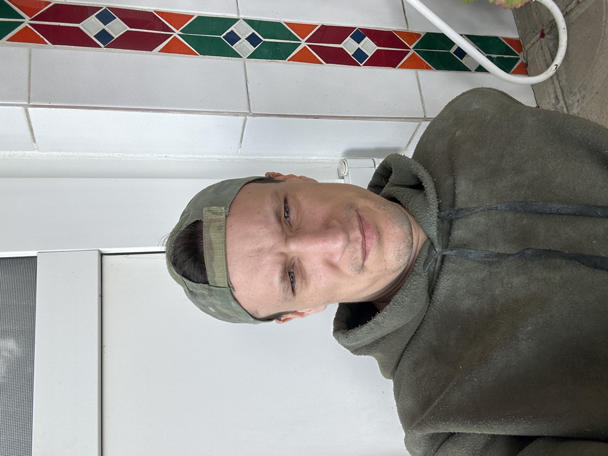 A man wearing a camouflage baseball cap and a dark green hooded sweatshirt is shown from the chest up. He is looking at the camera with a slight frown, his right eye squinting. The man has short dark hair and stubble on his face. Behind him, a white wall is visible, with a vertical decorative tile border to his right. The tile border features a geometric pattern in red, orange, green, and blue. A white pipe or railing is also visible in the lower right corner. The man appears to be standing near a doorway. The scene is likely outdoors, possibly on a porch or patio in the city of Makiv, Ukraine.