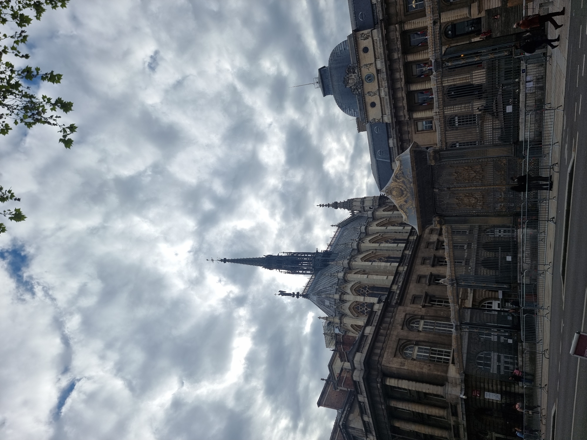 The image shows a building in Paris, France, likely the Conciergerie or Sainte-Chapelle based on its gothic architectural style. The buildings are constructed of stone, with intricate carvings and arched windows. A tall spire reaches toward the sky. Metal barriers line the street, separating the building from the public. A few people can be seen walking along the sidewalk. The sky is cloudy.