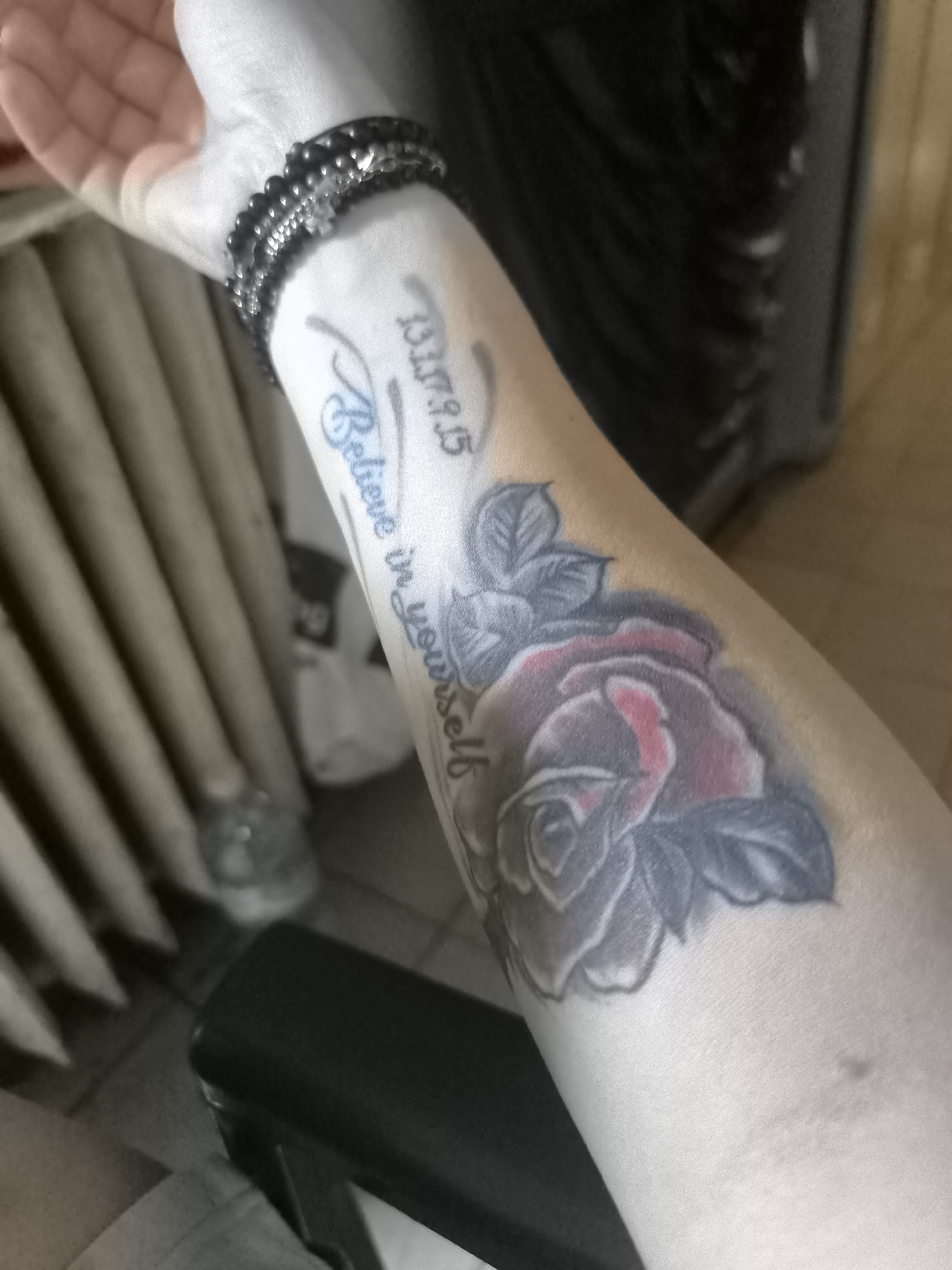 The image shows a close-up of a person's forearm with tattoos and a bracelet. The tattoos include a rose with dark grey petals and red accents, and text in blue and black ink. The blue text appears to read "Believe in yourself". Above this, black text includes the date "13.07.15". A black beaded bracelet with a silver charm is worn around the wrist. The forearm is resting against a ribbed, beige surface, possibly a radiator, on the left. In the background, a dark object, possibly furniture or clothing, is visible. The overall lighting suggests an indoor setting. The location of Sofia, Bulgaria, is not depicted in the image.
