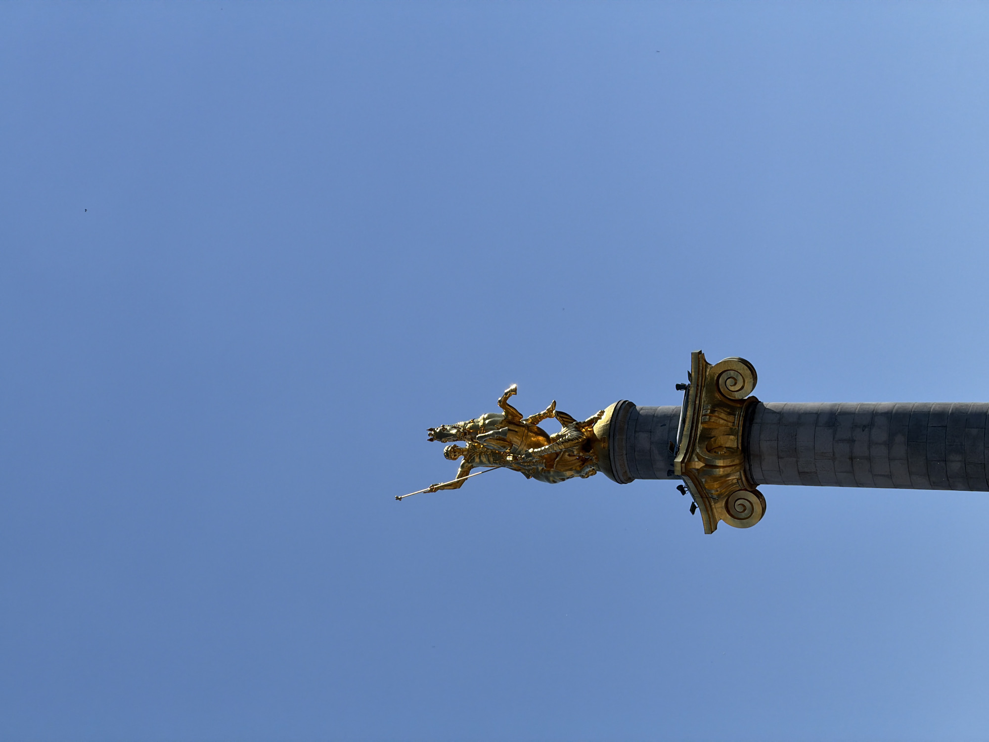 The image depicts a low-angle view of the Saint George statue atop the Freedom Monument in Tbilisi, Georgia, set against a clear blue sky. The statue is gold and depicts Saint George on horseback slaying a dragon. The statue is mounted on a grey stone column featuring a gold capital.