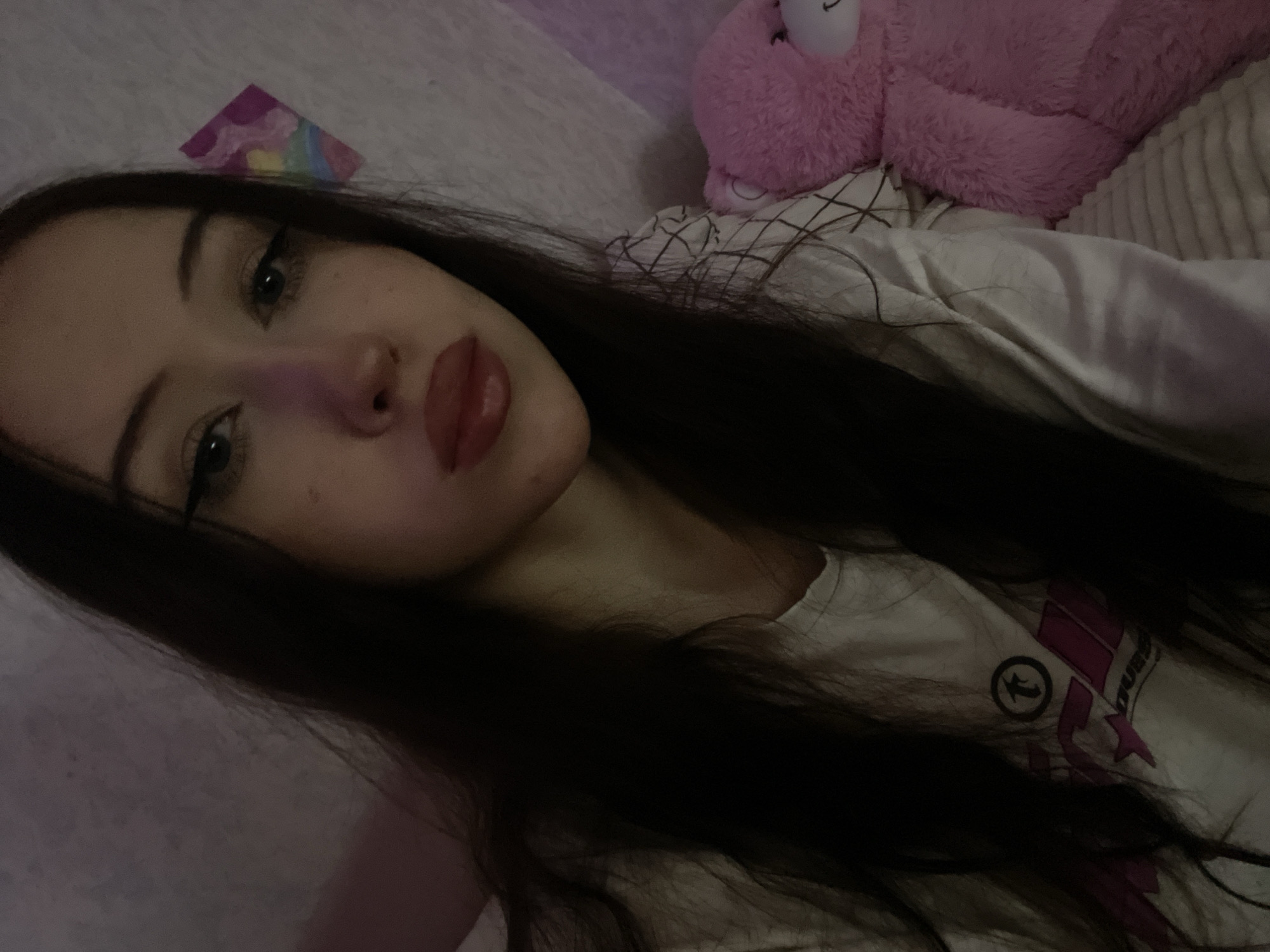 A young woman with dark hair is taking a selfie. She is wearing a white t-shirt with a graphic design and has makeup on her eyes and lips. To her right, a large pink teddy bear is visible. The background appears to be a wall with a small, colorful sticker. The overall lighting suggests an indoor setting, possibly a bedroom, with a pinkish hue. There is no indication of the specific location within the city of Votkinsk, Russia.