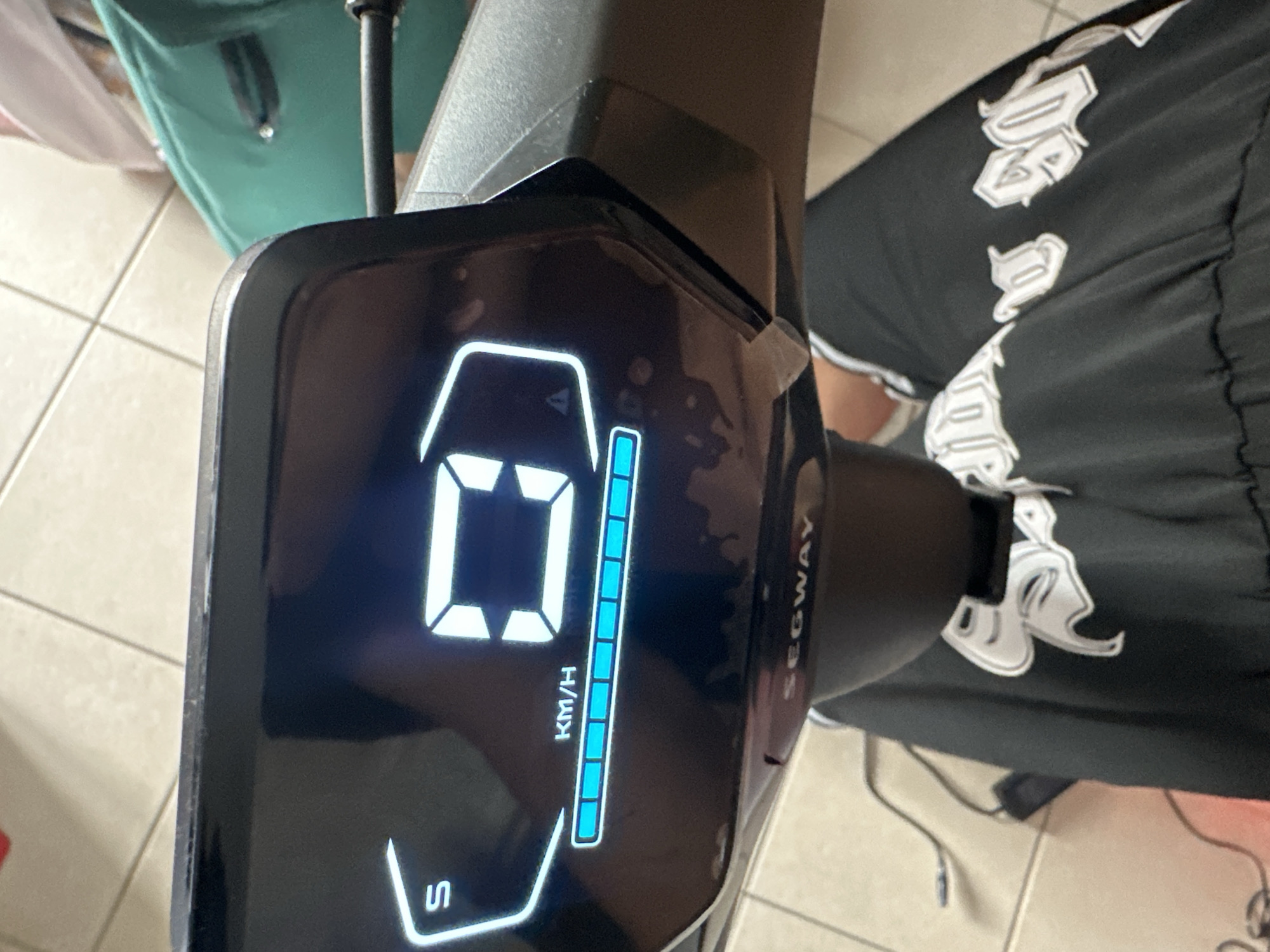The image shows a close-up of a digital display on a Segway Ninebot. The display shows a battery indicator with several blue bars filled, a speed reading of "0" km/h, and the letter "S" which likely indicates Sport mode. The Segway is black and has a logo that appears to be stylized white lettering. The background is blurred but appears to be an indoor setting with tiled flooring. There is no indication that this image is located in Anduze, France.