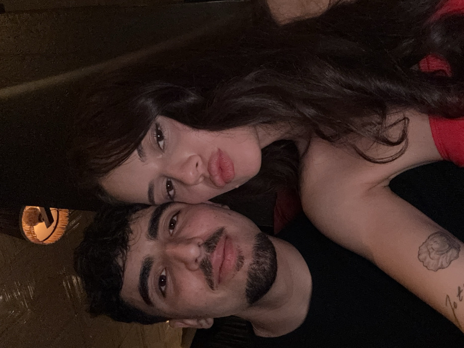 A brunette woman in a red top poses closely with a man wearing a black shirt. She has her head tilted, looking at the camera with pursed lips. The man also faces the camera, displaying a short beard and mustache. A tattoo is visible on the woman's arm. The image is slightly rotated, with dim lighting.