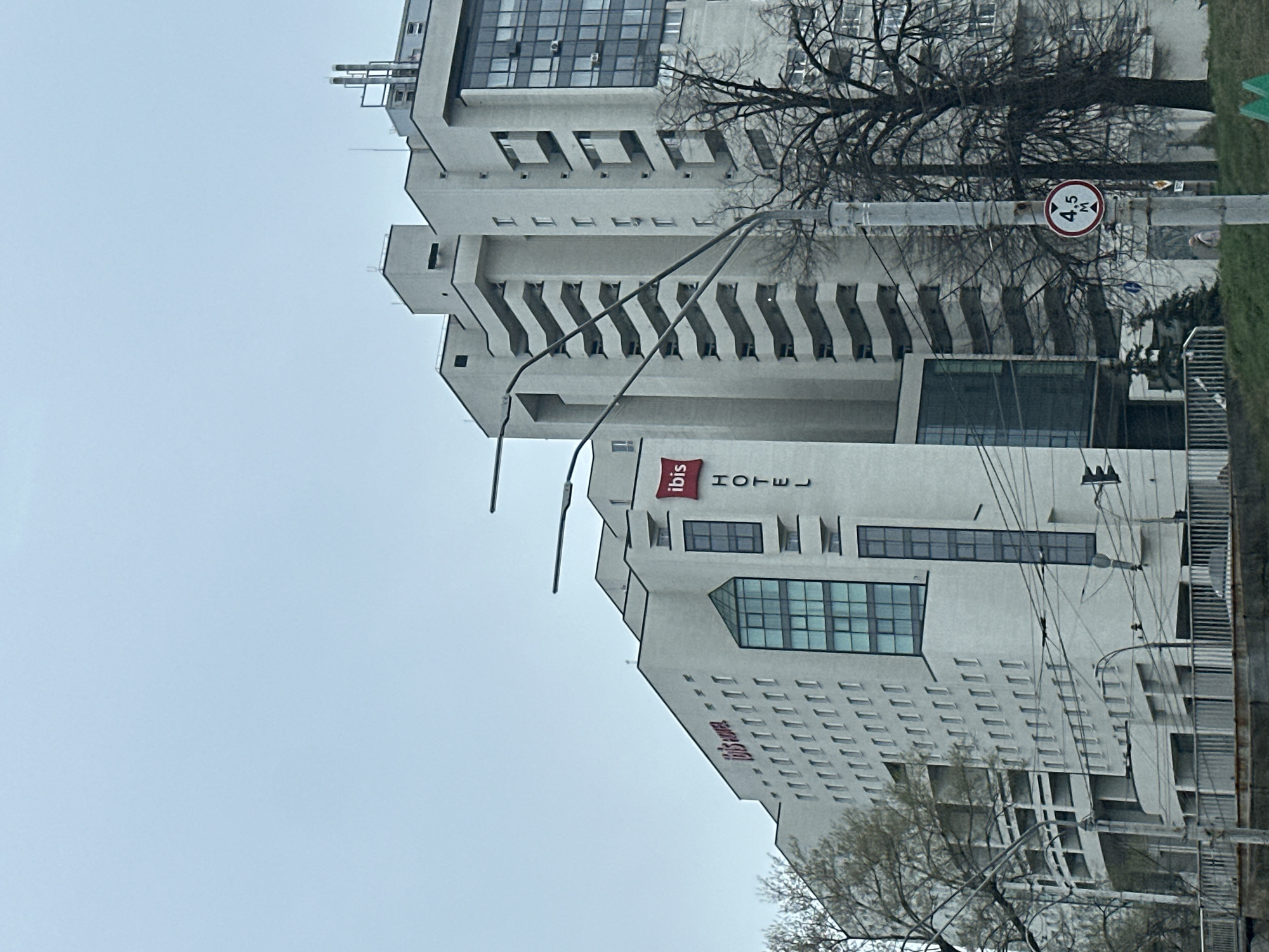 The image shows the Ibis hotel in Kyiv, Ukraine. The hotel is a tall, white building with multiple floors and a modern architectural style. The "Ibis HOTEL" signage is visible on the building's facade. A traffic sign indicating a height restriction of 4.5 meters is also present near the hotel. The sky is overcast, suggesting a cloudy day. There are trees with bare branches visible around the building, indicating that the season might be late autumn or winter. Utility poles and wires are also present in the foreground.