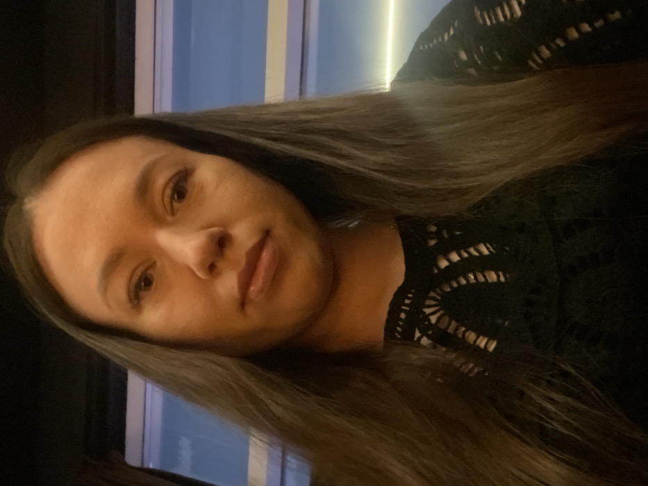 A medium-close-up photograph depicts an adult female looking directly at the camera. She has long, dark brown hair, centrally parted, extending over her shoulders. Her attire consists of a black top featuring an open-knit or crocheted pattern across the upper chest and neckline. The subject exhibits a neutral facial expression. The background reveals a window on the right side, through which a soft blue light and a distinct horizontal bright line are visible, suggesting an exterior during low-light conditions. The immediate environment appears indoors and dimly lit. No visual cues within the image identify a specific location in Perm, Russia.