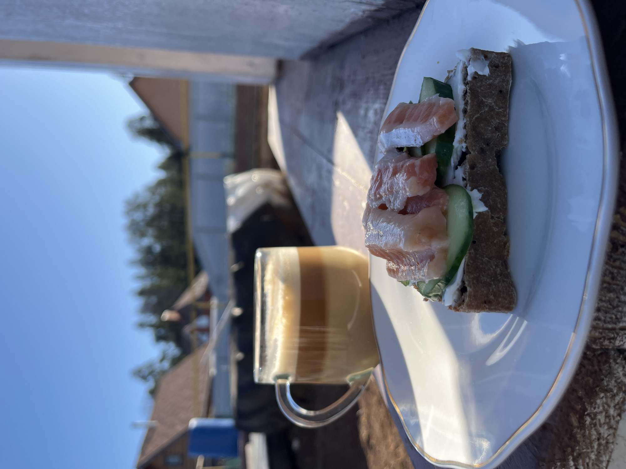 A close-up, angled, outdoor shot shows a sandwich and a layered beverage on a plate, appearing to be on a wooden surface.
The sandwich, which is on the right side of the frame, is placed vertically on a white plate with a gold rim. It consists of a dark bread base, spread with a white cream cheese, topped with slices of cucumber and what appears to be raw salmon.
Next to the sandwich, on the left side of the frame, is a clear glass mug with a layered beverage. The beverage appears to have a gradient from dark brown on the bottom to a lighter tan or cream color on top.
In the background, slightly out of focus, is a view of rooftops and trees against a clear blue sky. The location is specified as Chaikovsky, Russia, but there are no distinguishing features visible to confirm this. No people are visible in the shot.