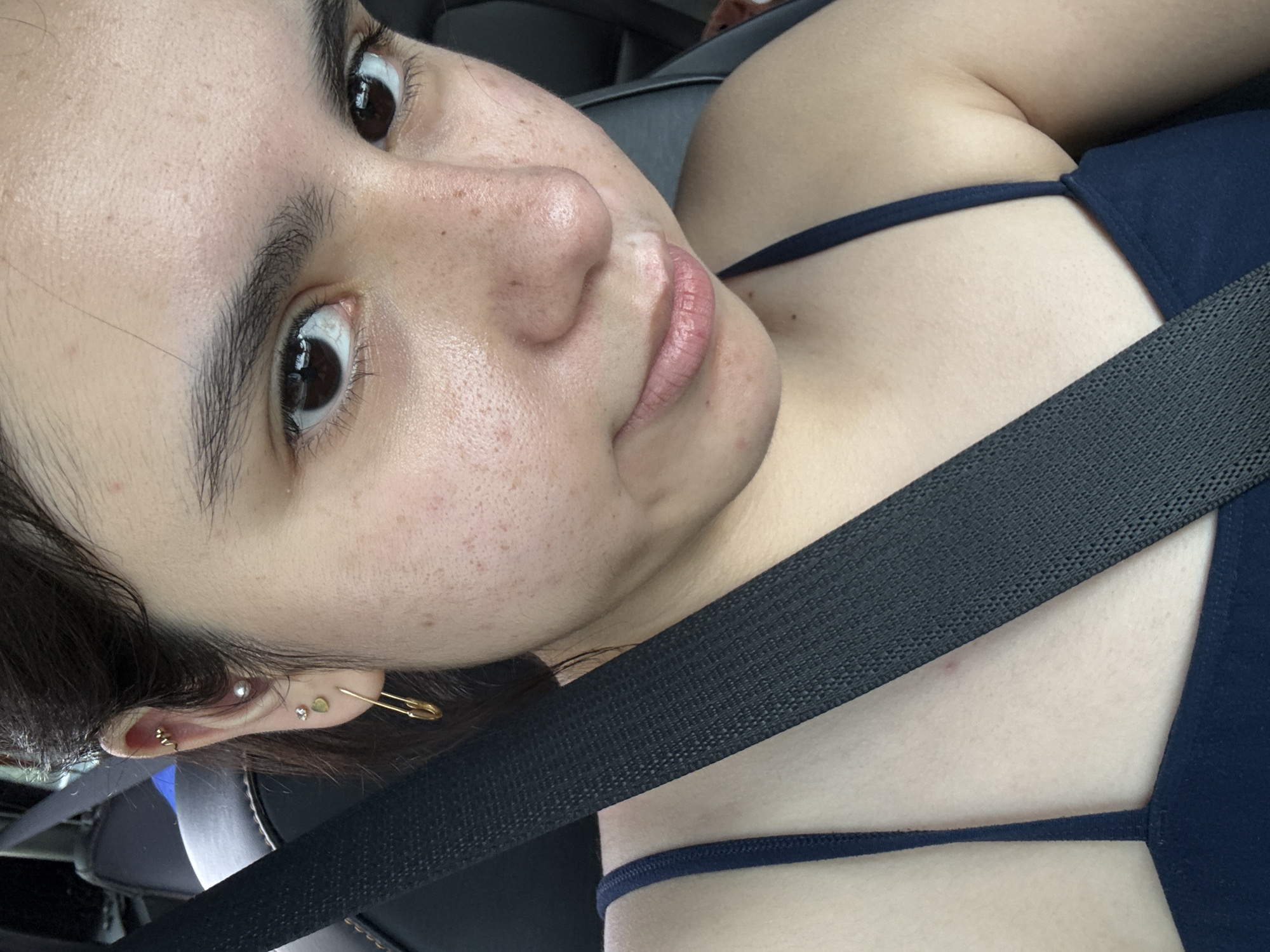 The image shows a person with fair skin and brown eyes wearing a dark blue top and a seatbelt in a vehicle. The person has multiple piercings in their left ear, including a safety pin. The background appears to be the interior of a car, with dark upholstery visible. The location cannot be determined from the image.