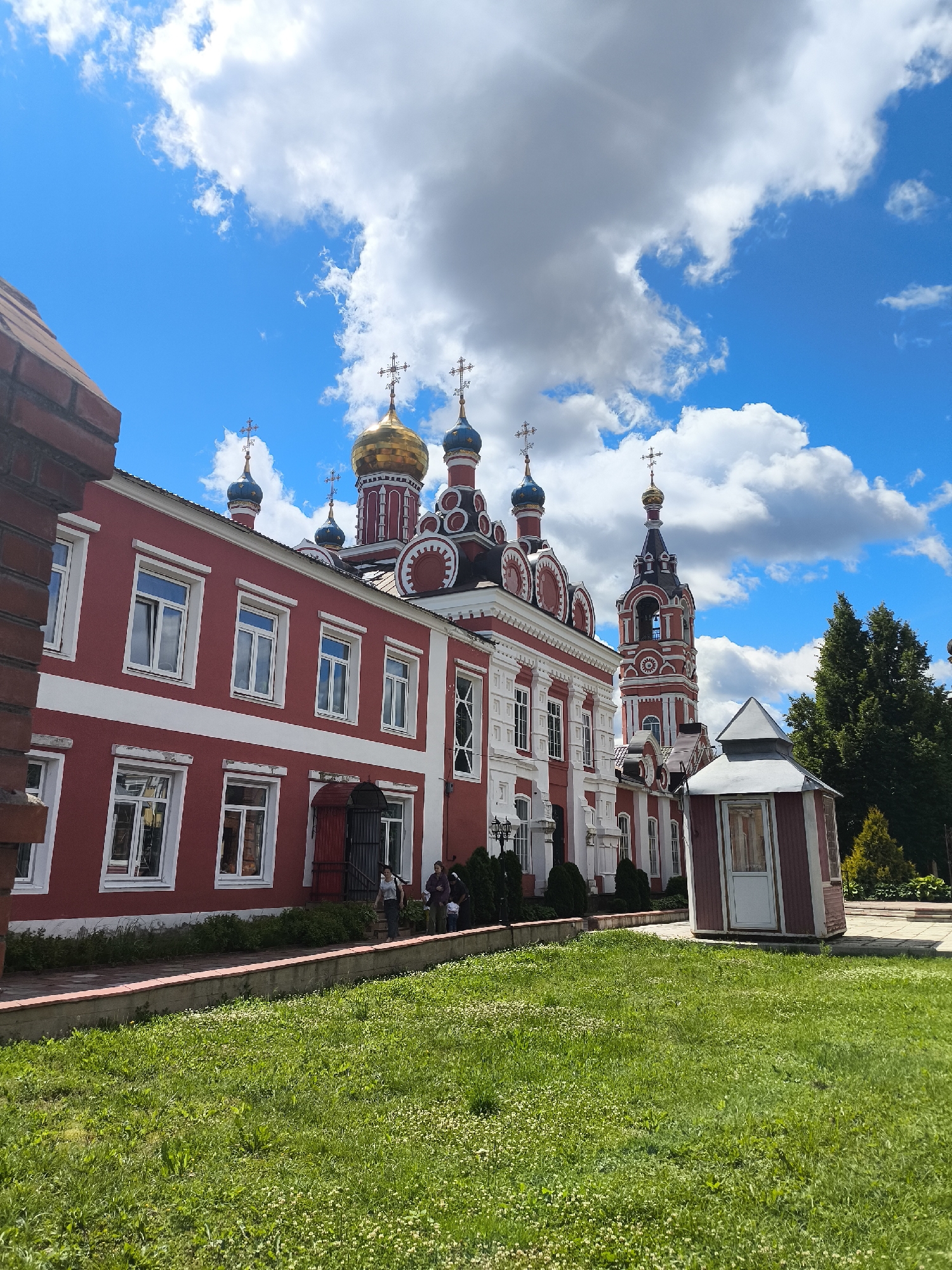 In the city of Taldom, Russia, a bright red building with white trim and multiple windows stands under a vivid blue sky dotted with white clouds. The building appears to be a church, with several onion domes topped with crosses visible in the background, including a prominent golden dome. A bell tower with red and white detailing is also present. In front of the building, a manicured green lawn slopes down towards the viewer. A small, red, shed-like structure with a white door sits to the right of the building. A few individuals are walking near the entrance of the red building.