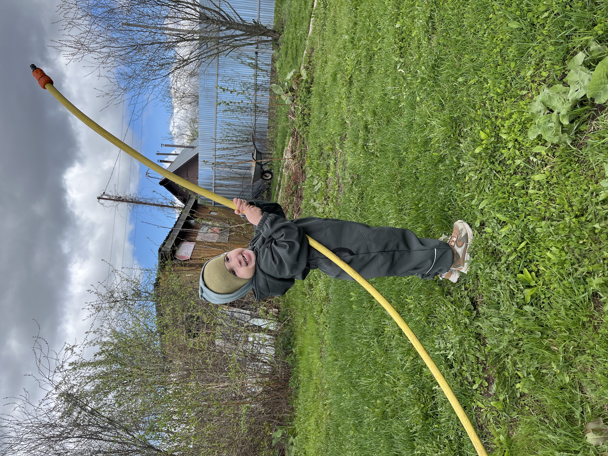 A child in a dark green jumpsuit and a light green hat is holding a yellow garden hose in an outdoor setting. The location is Chaikovsky, Russia. The hose curves upward and away from the child, who is smiling and appears to be playing with it. The setting includes a lawn with green grass, trees with bare branches, a fence made of corrugated metal, and a small wooden structure that could be a shed or part of a house. The sky is partly cloudy.