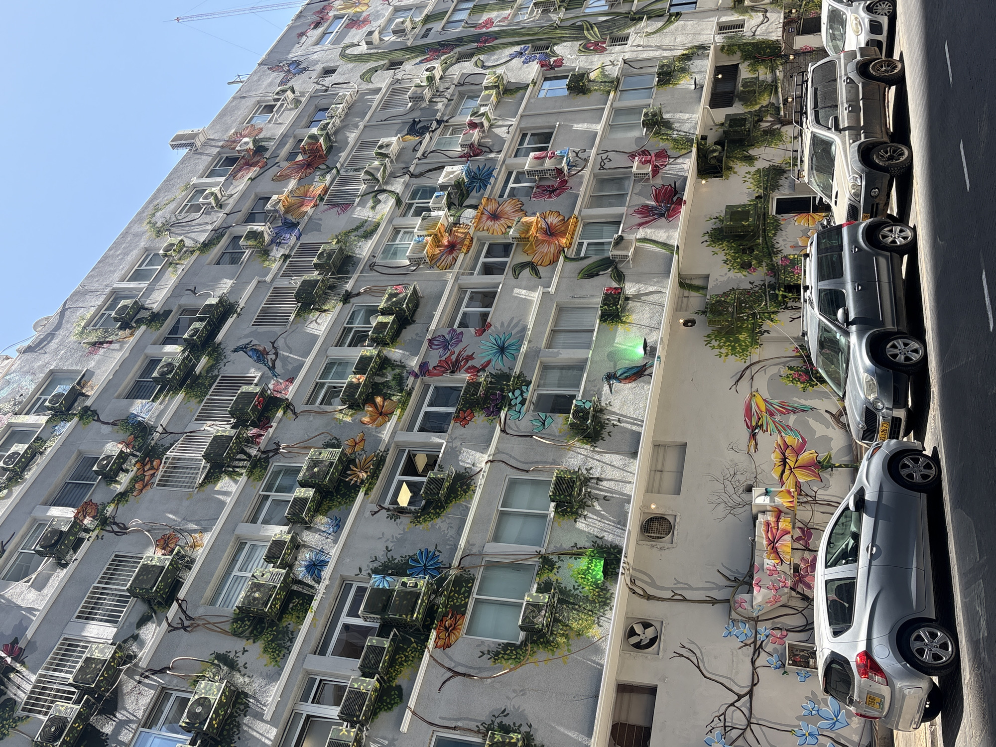 The image displays a tall, multi-story building in Tel-Aviv, Israel, covered by an extensive and colorful mural. The building's facade, rendered in a light grey hue, is adorned with an elaborate botanical design featuring large, vibrant flowers in shades of orange, yellow, red, blue, and purple, interspersed with green vines, leaves, butterflies, and birds. Numerous air conditioning units are mounted on the building, many of which are cleverly integrated into the mural by being painted with matching leafy camouflage patterns.
The building is oriented diagonally in the frame, creating a dynamic perspective against a clear blue sky. Bright sunlight illuminates the scene, causing luminous green reflections on some windows and air conditioning units. In the upper left corner, a portion of a construction crane is faintly visible against the sky.
Along the right side of the building, several cars are parked on a paved street. These include a light grey hatchback, a larger dark grey SUV-like vehicle with a roof rack, and another light-colored car further down the street. White painted lines are visible on the road surface.
The scene depicts a sunny daytime environment with clear weather. No people are visible, and there is no explicit activity taking place beyond the static urban landscape enhanced by the mural. A partial license plate, "6142673", is visible on one of the parked cars.