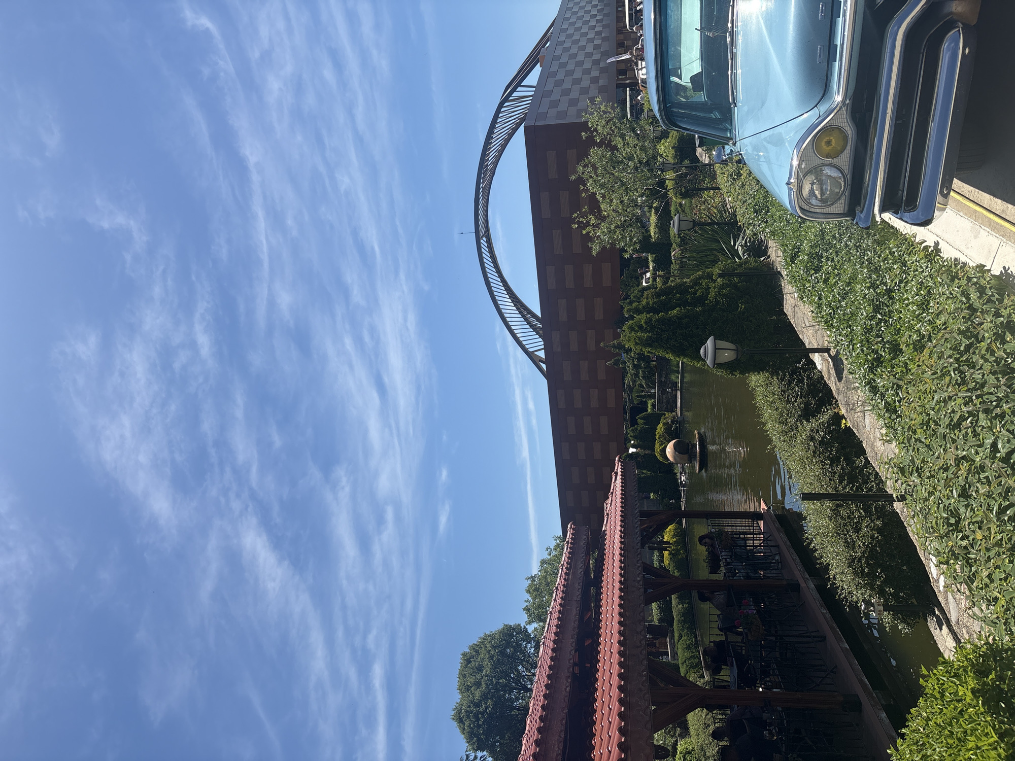 The image presents a scene of urban architecture and natural elements under a bright, clear sky with scattered clouds. A blue vintage car is partially visible in the foreground on the right side of the image. Adjacent to the car, a stone wall is covered with dense green bushes. Behind the wall is a serene pond with a fountain in the center. A wooden gazebo structure with a red tile roof is situated along the edge of the pond. Behind the pond rises a modern architectural structure consisting of a large building with a distinctive arch, built from brown concrete blocks. The photo is taken from a low angle, emphasizing the height and design of the building and the expansive blue sky.