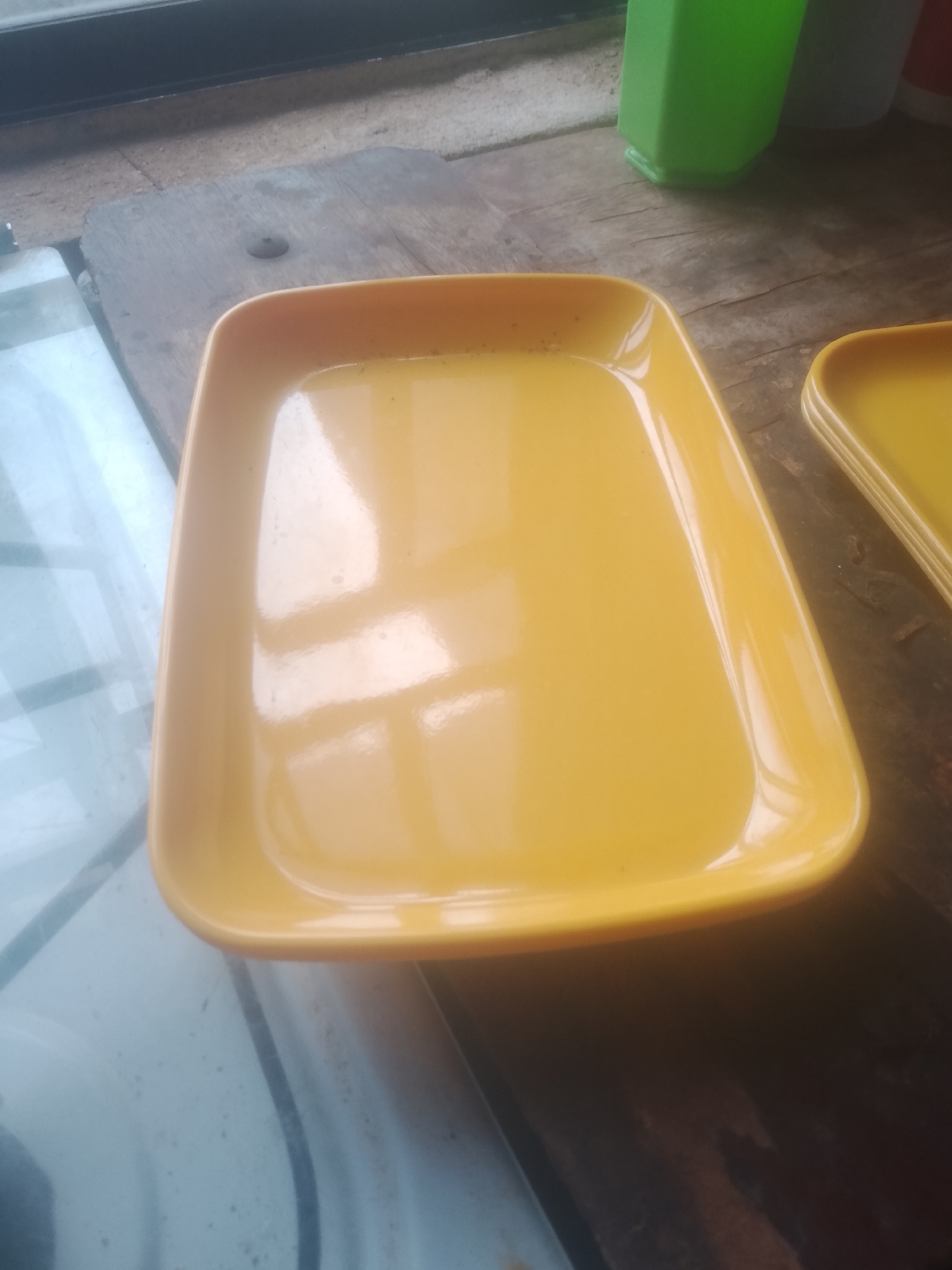 A close-up, slightly angled shot shows a yellow rectangular plate with rounded corners. The plate is empty and appears to be made of plastic or melamine. It is positioned on a rough wooden surface, possibly a table or shelf. To the right of the main plate, another identical yellow plate is visible, stacked slightly beneath it. In the background, a portion of a green plastic container or jug can be seen, along with what appears to be a window frame and a white surface that could be part of a refrigerator or other appliance. The lighting suggests an indoor setting, possibly a kitchen or dining area. The overall scene is static, with no visible human presence or activity. The location is specified as Jalingo, Nigeria.