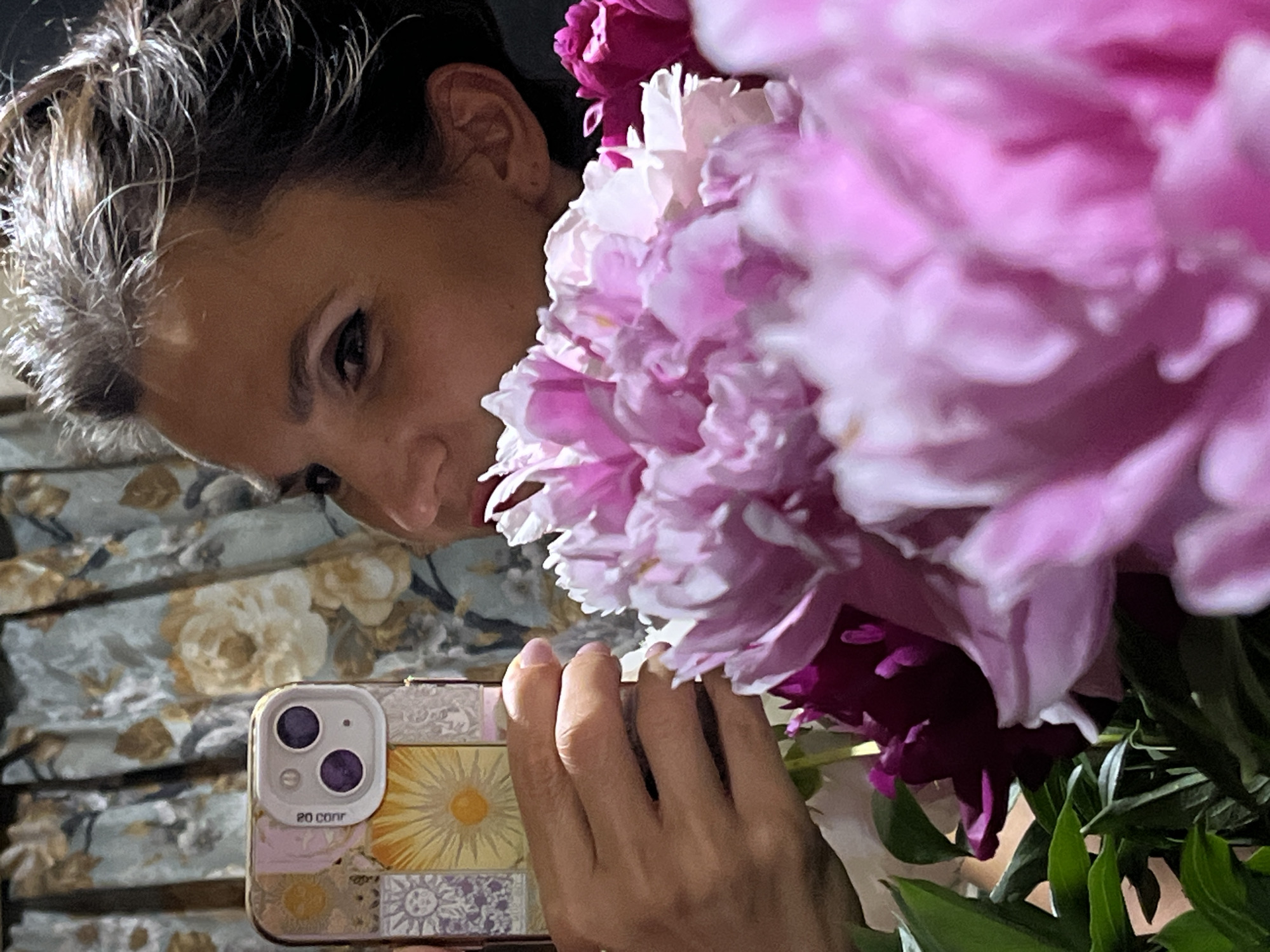A woman is taking a selfie with a bouquet of pink peonies in front of a floral curtain. She is holding her iPhone in her left hand, and the phone case has a collage of suns and celestial motifs. The woman's face is partially visible, with one eye looking towards the camera. The peonies are in full bloom and fill the foreground of the image. The background is a curtain with a light blue and cream floral pattern. The image appears to be taken indoors. There is no information in the image to suggest a specific location in Petropavlovsk-Kamchatsky, Russia.