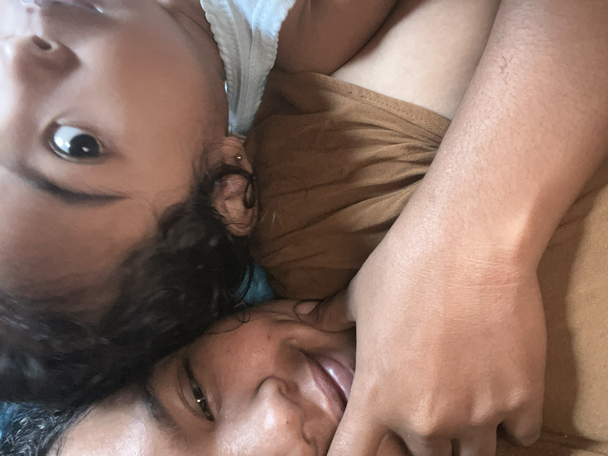 Two people are nestled together, with one person's head resting on the other's shoulder. The person whose head is on the shoulder has their eyes wide open, looking up and to the left. Their face is very close to the camera. The other person's face is partially visible, with their eyes closed and a slight smile. Both individuals have dark hair and light brown skin. They appear to be indoors, in a comfortable setting, possibly a bed or couch, given the soft textures of their clothing. The lighting is warm and natural, suggesting it may be daytime. The image is a close-up shot, focusing on the faces and upper bodies of the two individuals. The overall impression is one of intimacy and affection. There are no discernable elements in the image that would suggest a specific location in Kruengmate, Indonesia.